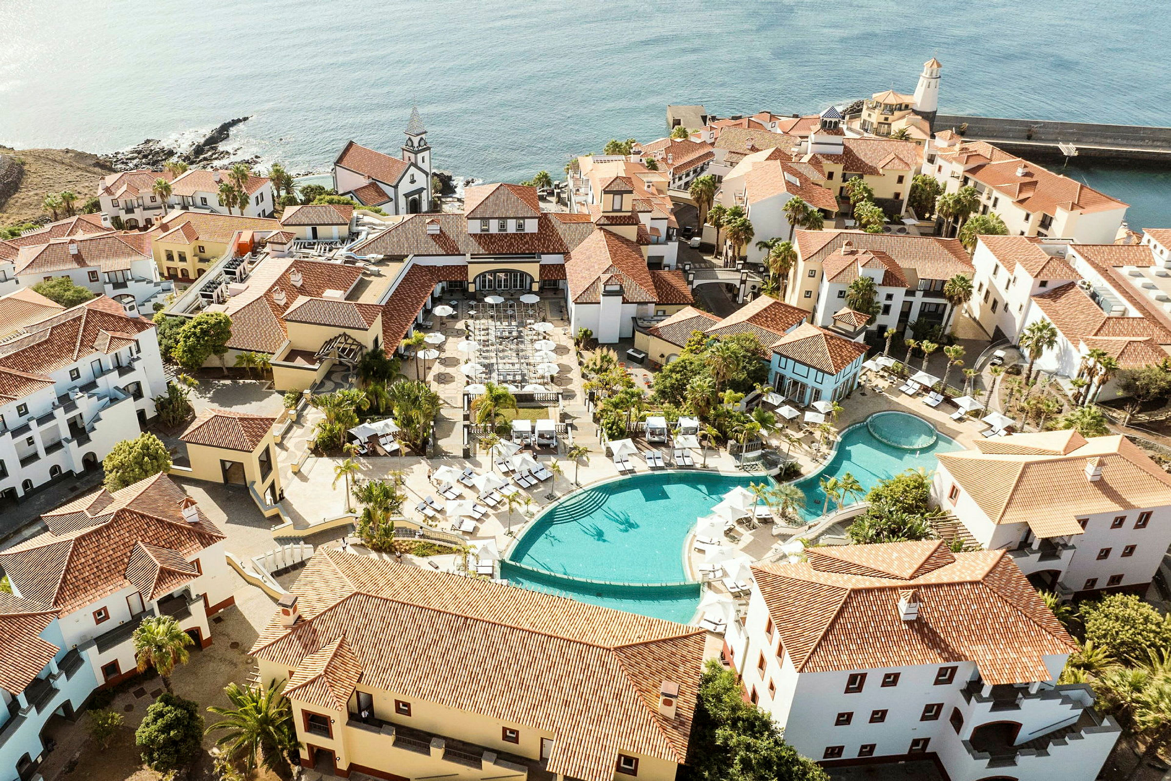 Luftfoto af et feriested ved havet med swimmingpools, liggestole og omkringliggende bygninger i middelhavsstil. (AI-genereret tekst)