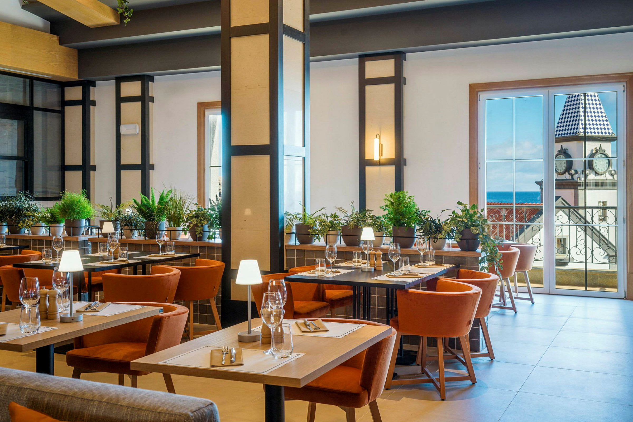 En velbelyst restaurantinteriør med borde, stole og grøn planteudsmykning. I baggrunden er der store vinduer, der viser udsigt over en bygning med et klokketårn. (AI-genereret tekst)