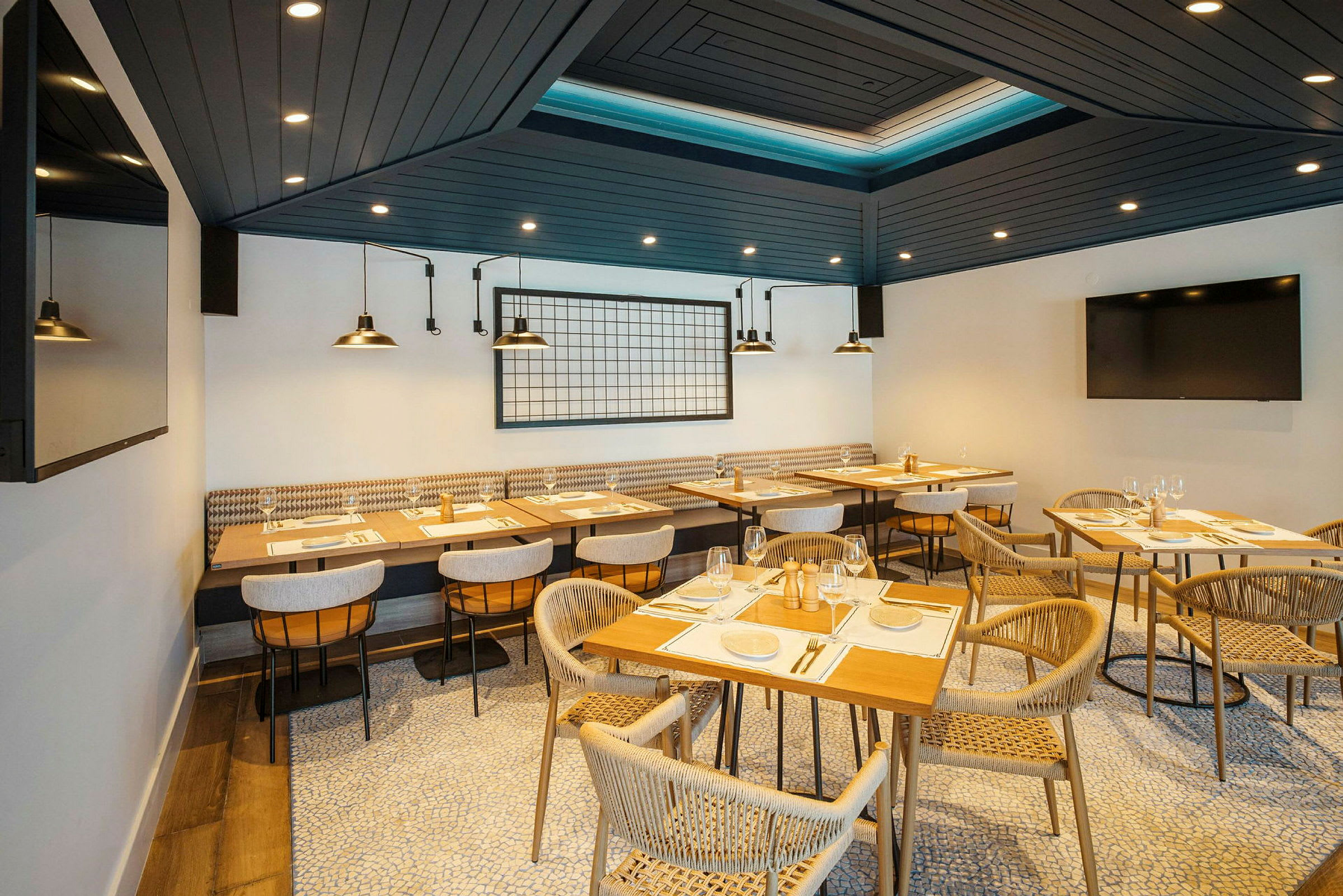 En moderne restaurant med træborde og komfortable stole arrangeret på et organisk mønstret gulv. Loftet er mørkt med spots, og bagvæggen har industrielle lamper og dekoration. (AI-genereret tekst)