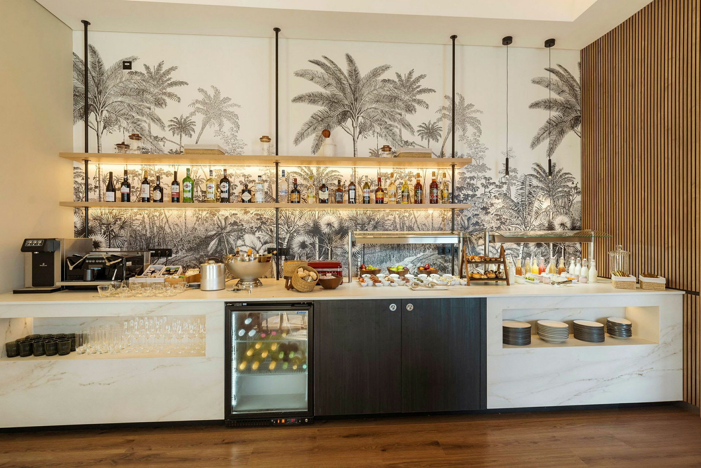 Billede af en morgenmadsbuffet på et hotel med forskellige drikkevarer, mad og serveringsudstyr samt en dekorativ væg med palmemønster i baggrunden. (AI-genereret tekst)