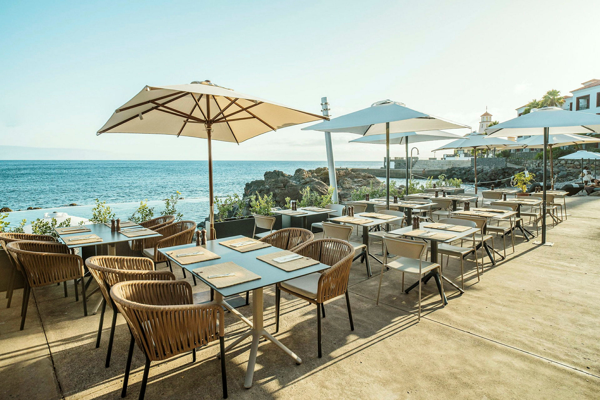 Udendørs restaurantområde med borde og stole under hvide parasoller med udsigt over havet og klipperne. (AI-genereret tekst)