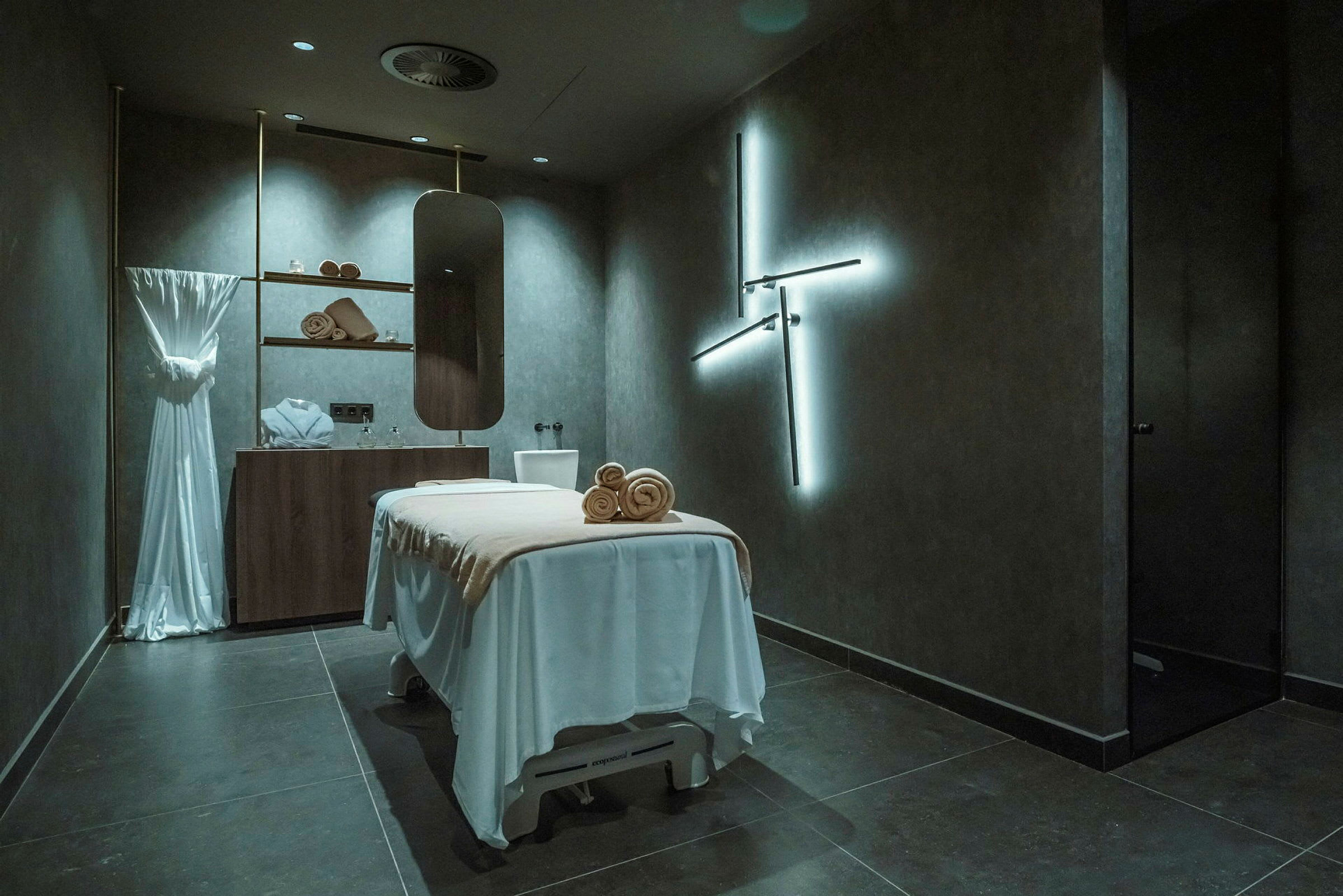Et rum med en massageseng dækket med hvidt, hylder med håndklæder og dekorationer samt et spejl på væggen. Rummet har en moderne og afslappende atmosfære. (AI-genereret tekst)