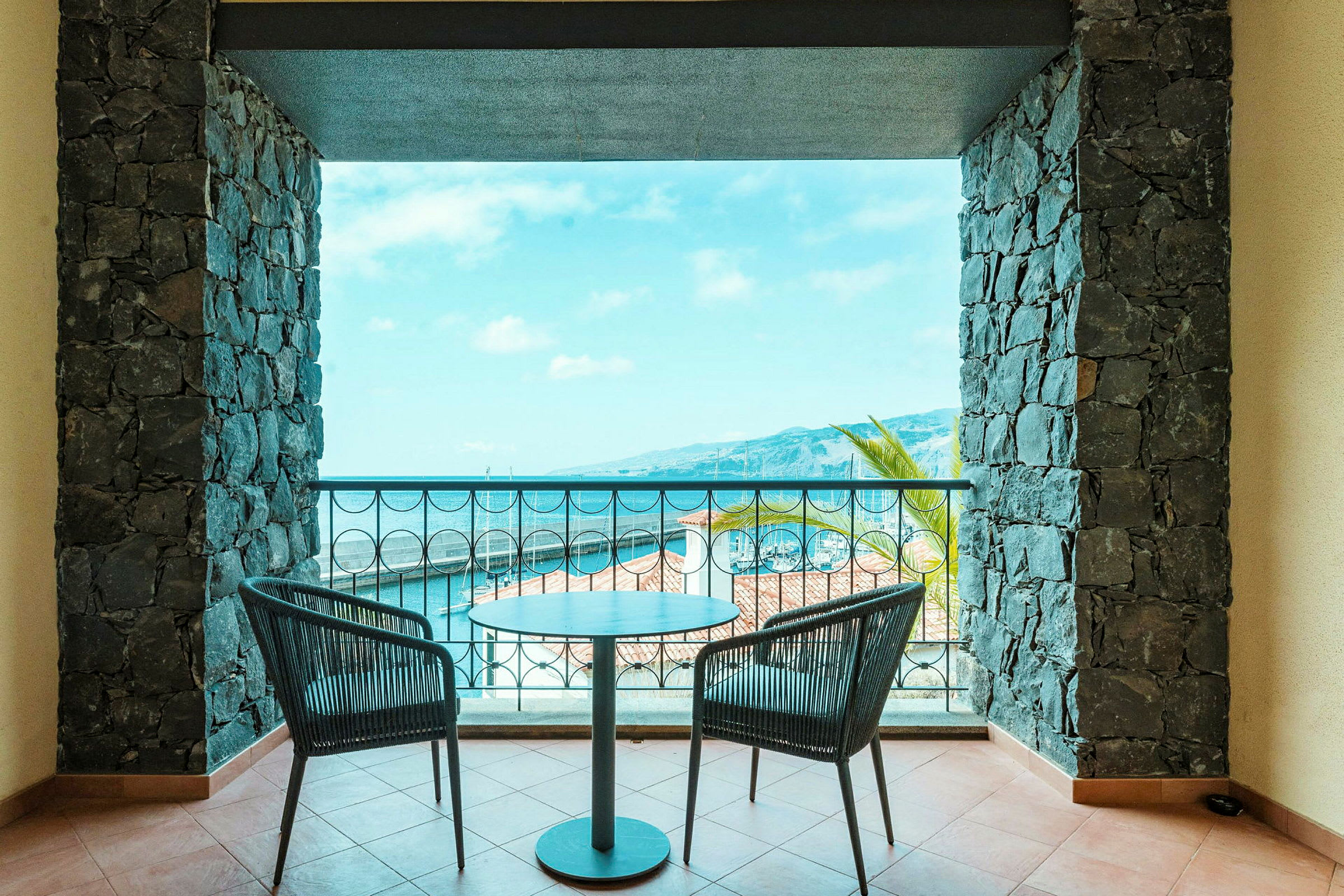 En balkon med to siddepladser og et bord, omgivet af stenvæg med udsigt over havet og marinaen. (AI-genereret tekst)
