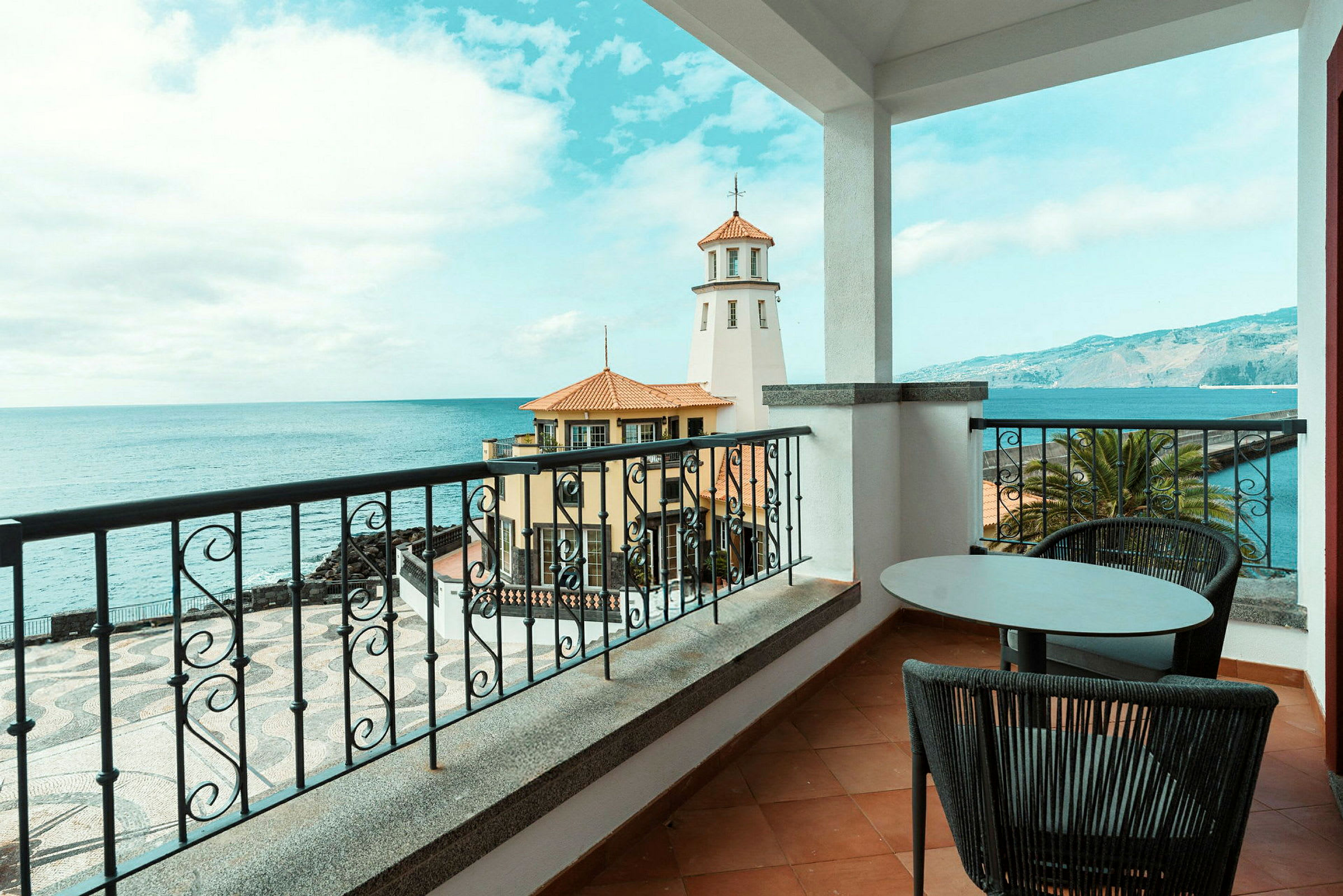 Udsigt fra en balkon mod havet med en fyrtårn og bygninger i forgrunden. (AI-genereret tekst)