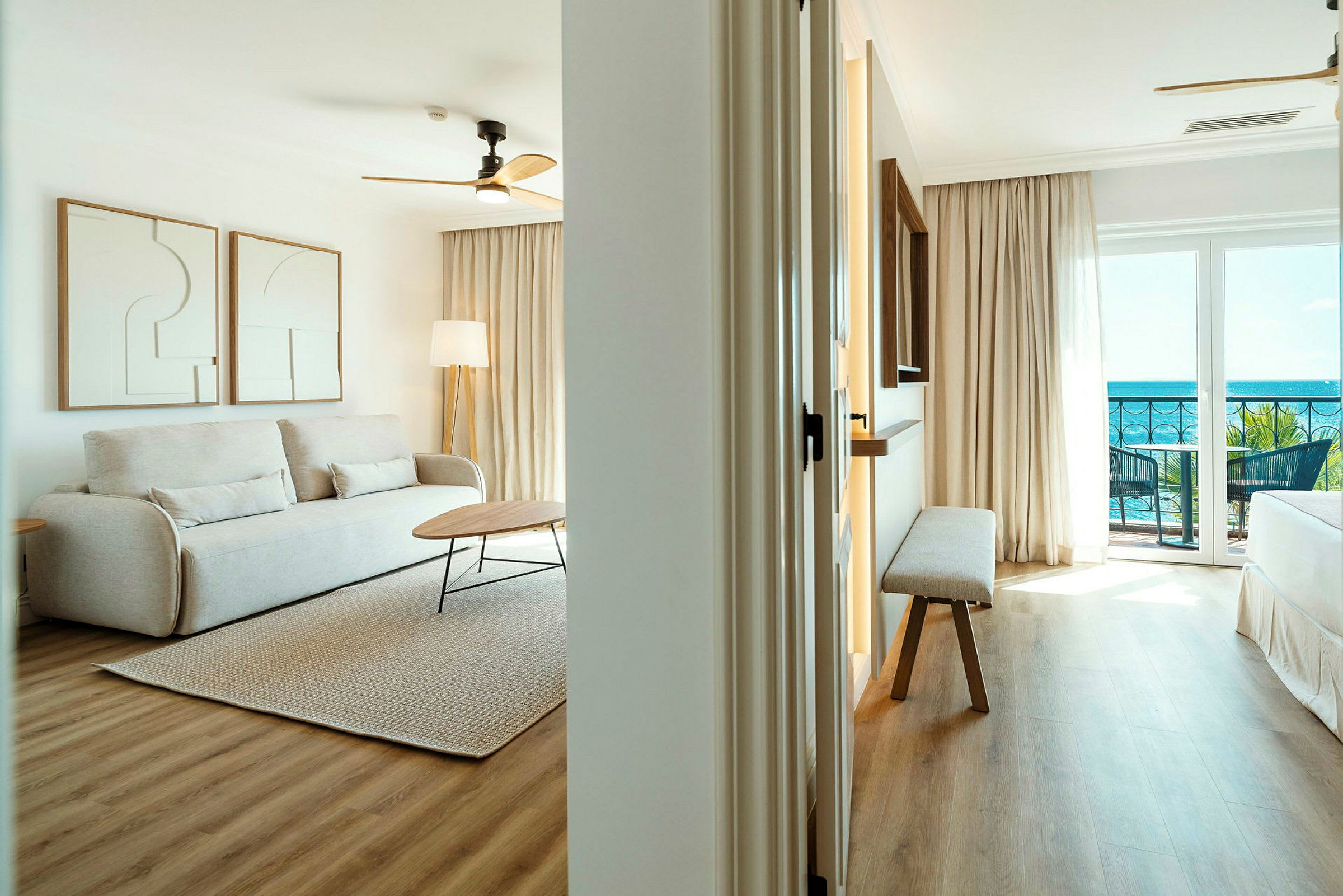 Et lyst opholdsområde med en beige sofa, to kunstværker på væggen, et bord og et tæppe på trægulv. Til højre anes et soveværelse med udsigt til havet. (AI-genereret tekst)
