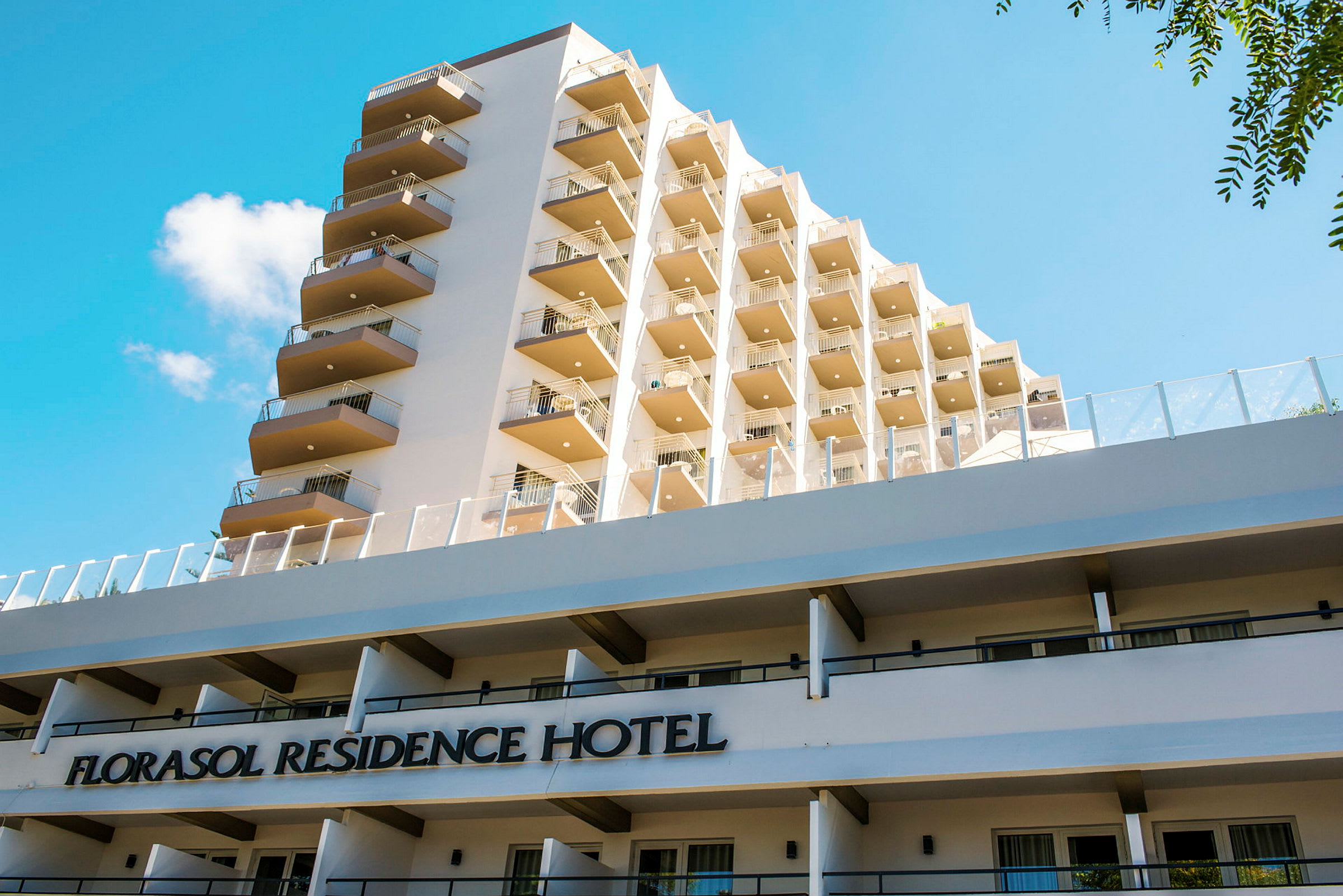 Udsigt over bygningen Florasol Residence Hotel med flere altaner og en blå himmel i baggrunden. (AI-genereret tekst)