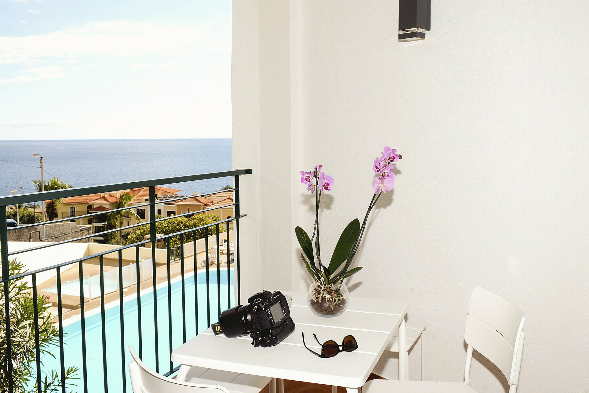 Udsigt fra en balkon med havet i baggrunden, en pool synlig nedenfor. På balkonen er der et bord med et kamera, solbriller og en orkidé. (AI-genereret tekst)
