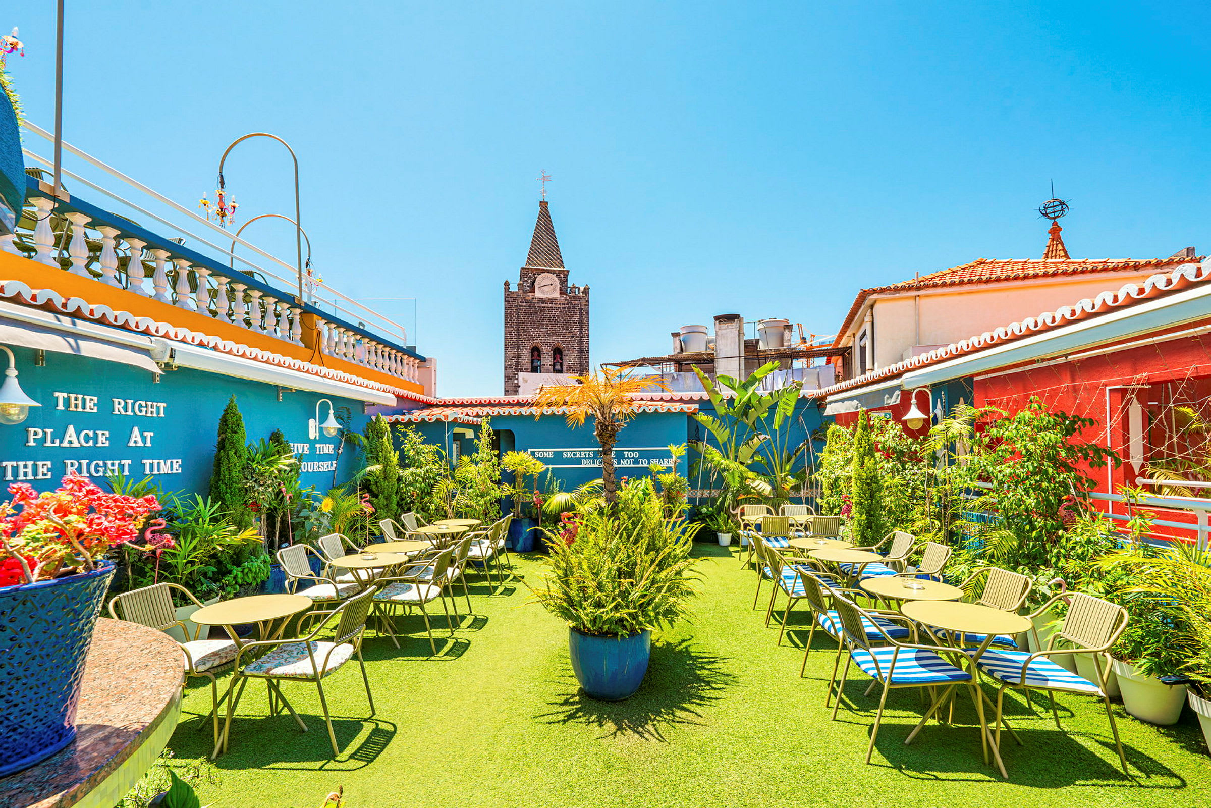 En tagterrasse med borde og stole, omgivet af grønne planter og farverige bygninger. (AI-genereret tekst)