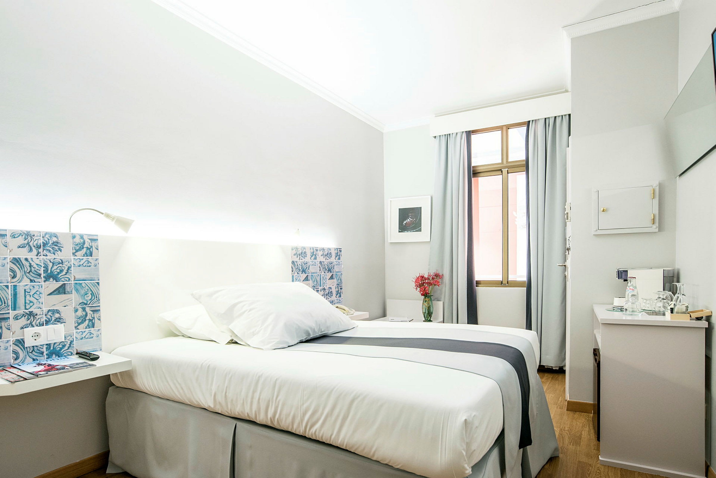 Et lyst og stilrent hotelværelse med en enkeltseng dækket med hvide sengelinned og et natbord ved siden. Vinduet har lysegrå gardiner. (AI-genereret tekst)