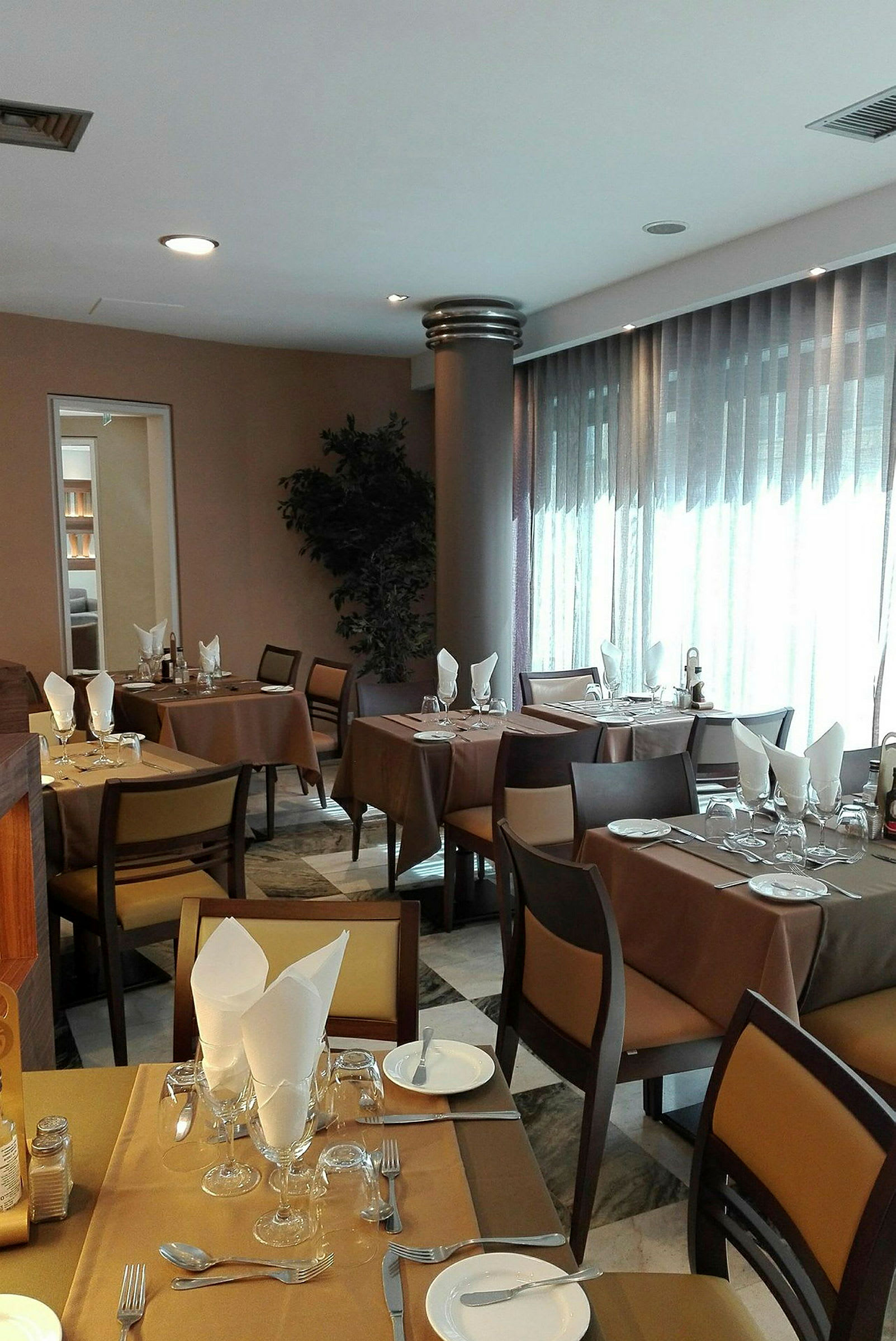 Et indendørs restaurantområde med dækkede borde, brune duge og stole med lysebrune puder. (AI-genereret tekst)