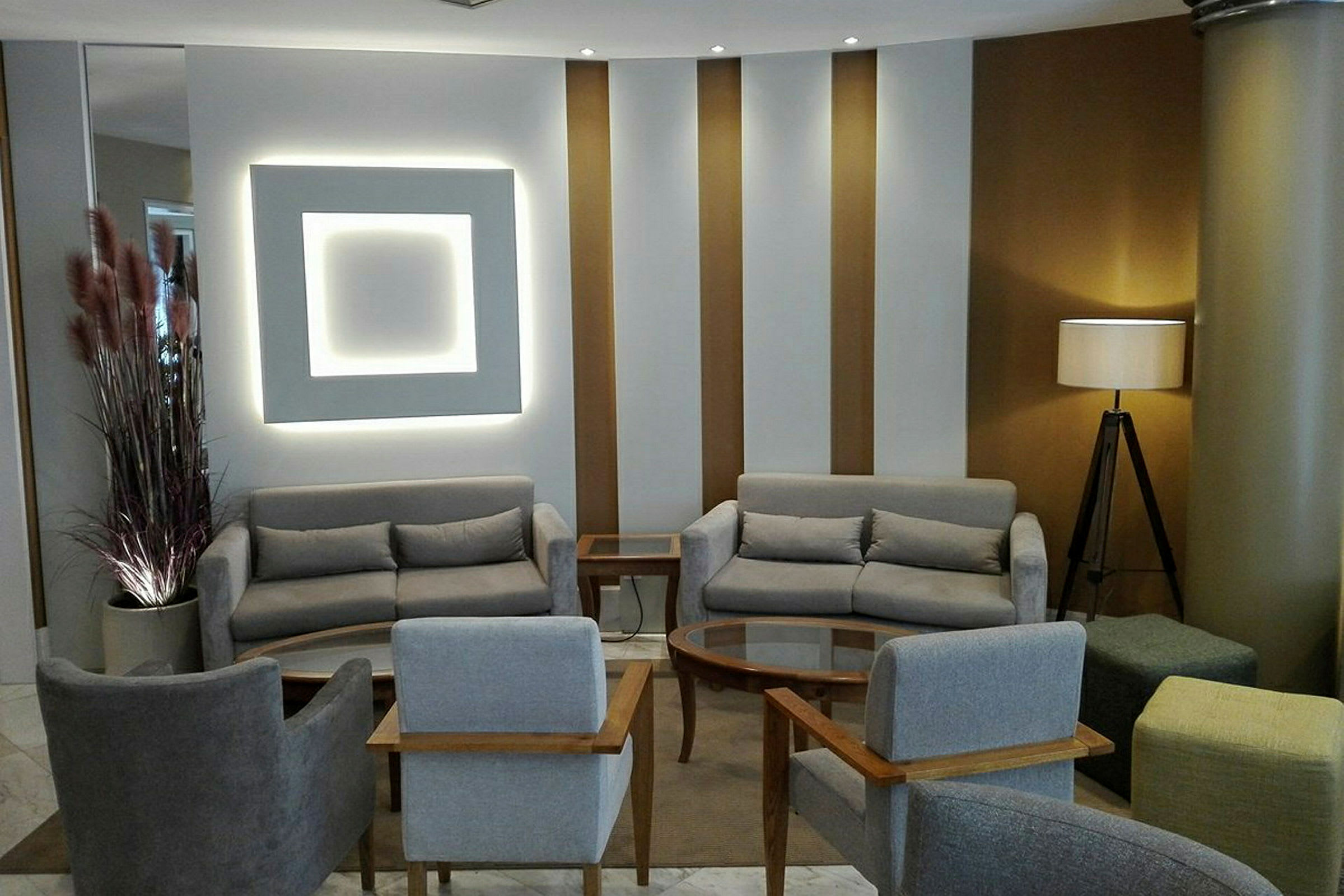 Et moderne loungeområde med grå sofaer og stole arrangeret omkring et sofabord på et tæppe. En lampe står i hjørnet ved en søjle. (AI-genereret tekst)