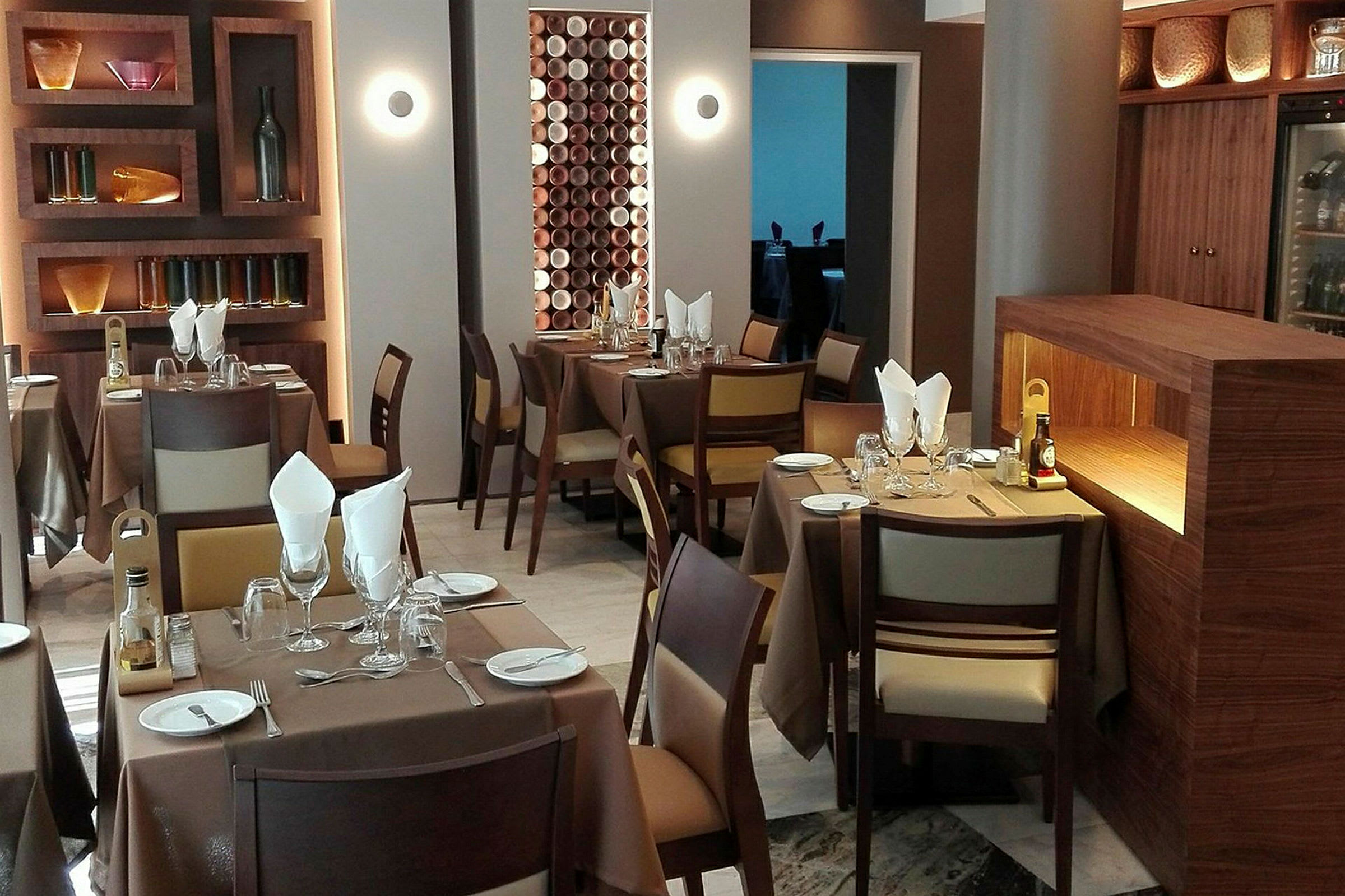 En moderne restaurant med borde dækket med brune duge og matchende træstole. I baggrunden er væghylder med dekorative genstande. (AI-genereret tekst)