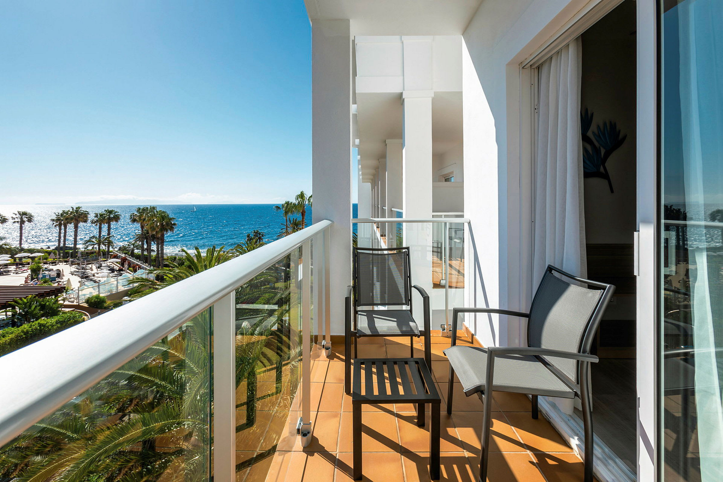 En balkon med to stole og et bord, med udsigt over palmer og havet. (AI-genereret tekst)