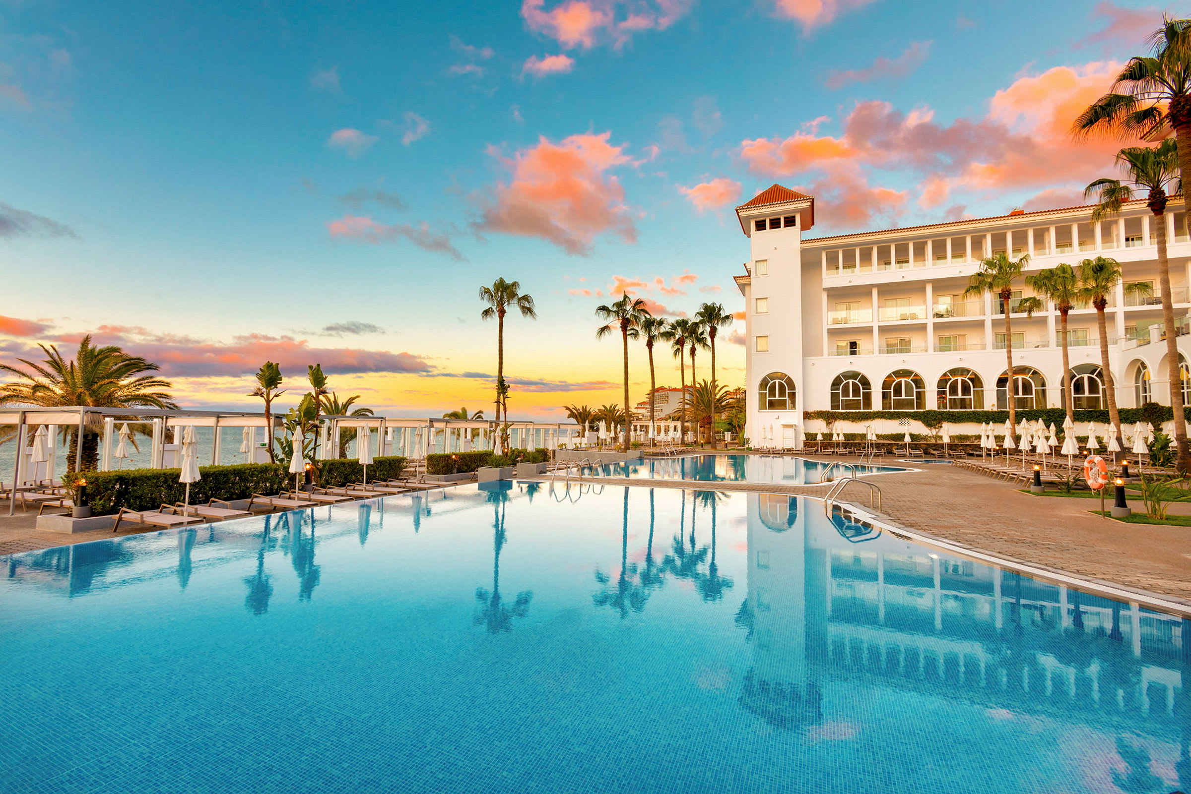 Hotel med store vinduer og pool, omgivet af liggestole og palmer ved solnedgang. (AI-genereret tekst)