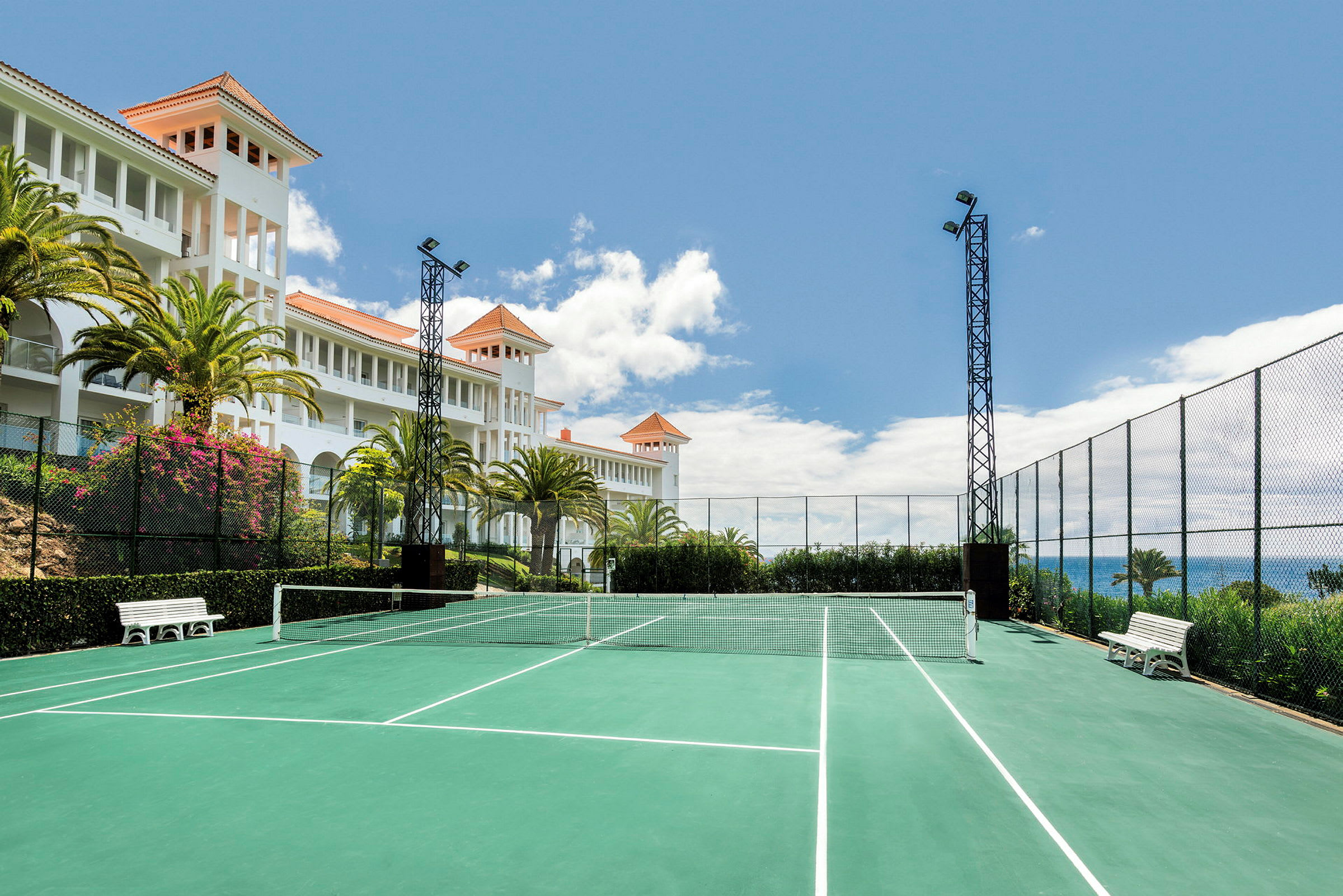 En tennisbane med grøn overflade og hvide linjemarkeringer er omgivet af hegn. I baggrunden ses en hvid bygning med orange tag og palmer samt en blå himmel. (AI-genereret tekst)