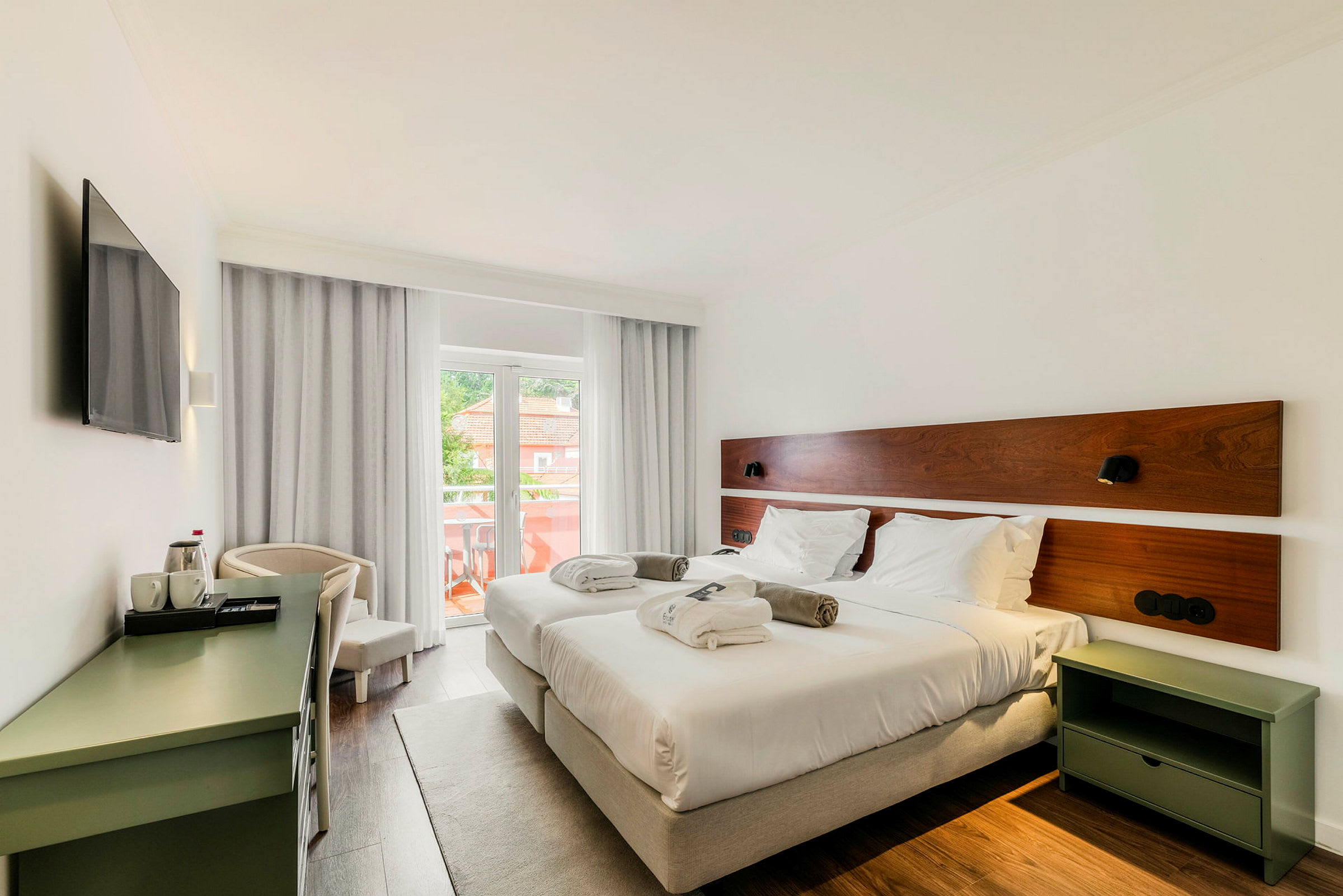 Et hotelværelse med to enkeltsenge, hvidt sengetøj og puder. Værelset har en balkon med udsigt og er indrettet med et skrivebord og et tv. (AI-genereret tekst)