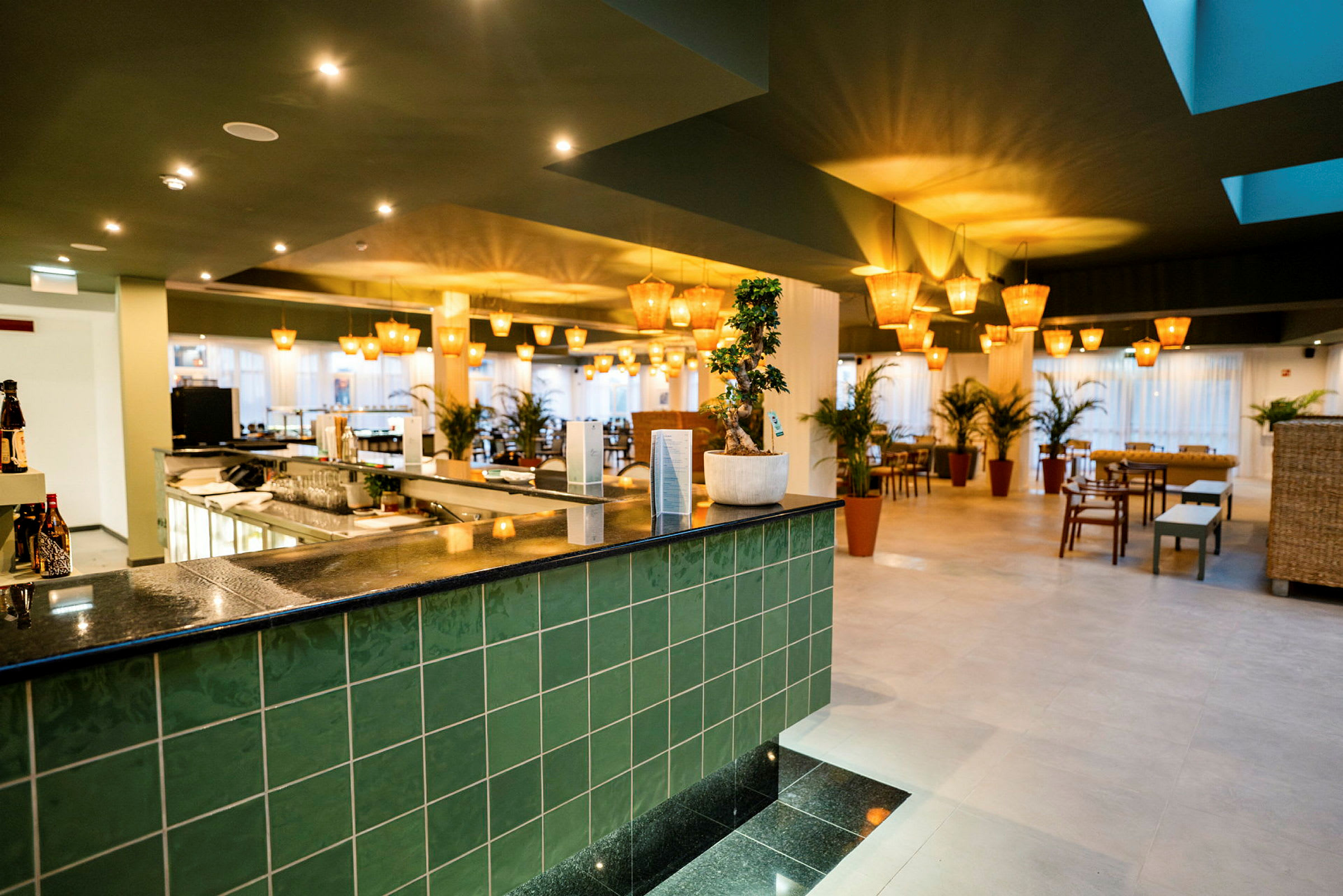 Et interiør af en moderne restaurant med grønne fliser, en bardisk og flere borde med stole samt planter. (AI-genereret tekst)