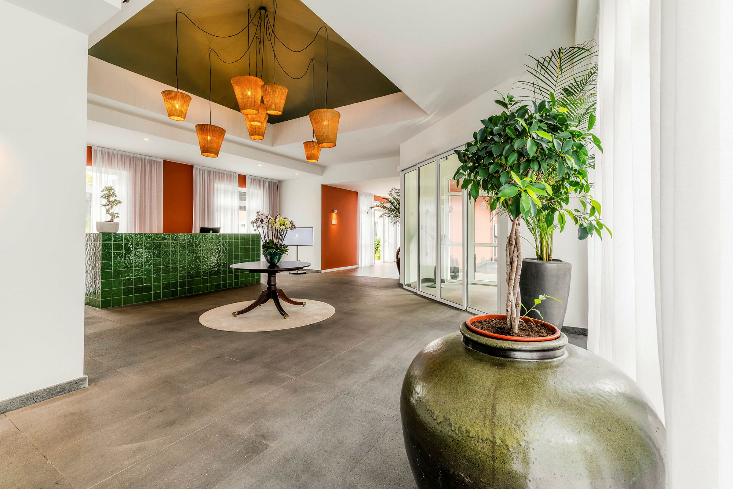 En lys og moderne hotellobby med grønne keramiske vægdetaljer, et rundt bord med blomster og store planter i forgrunden. (AI-genereret tekst)