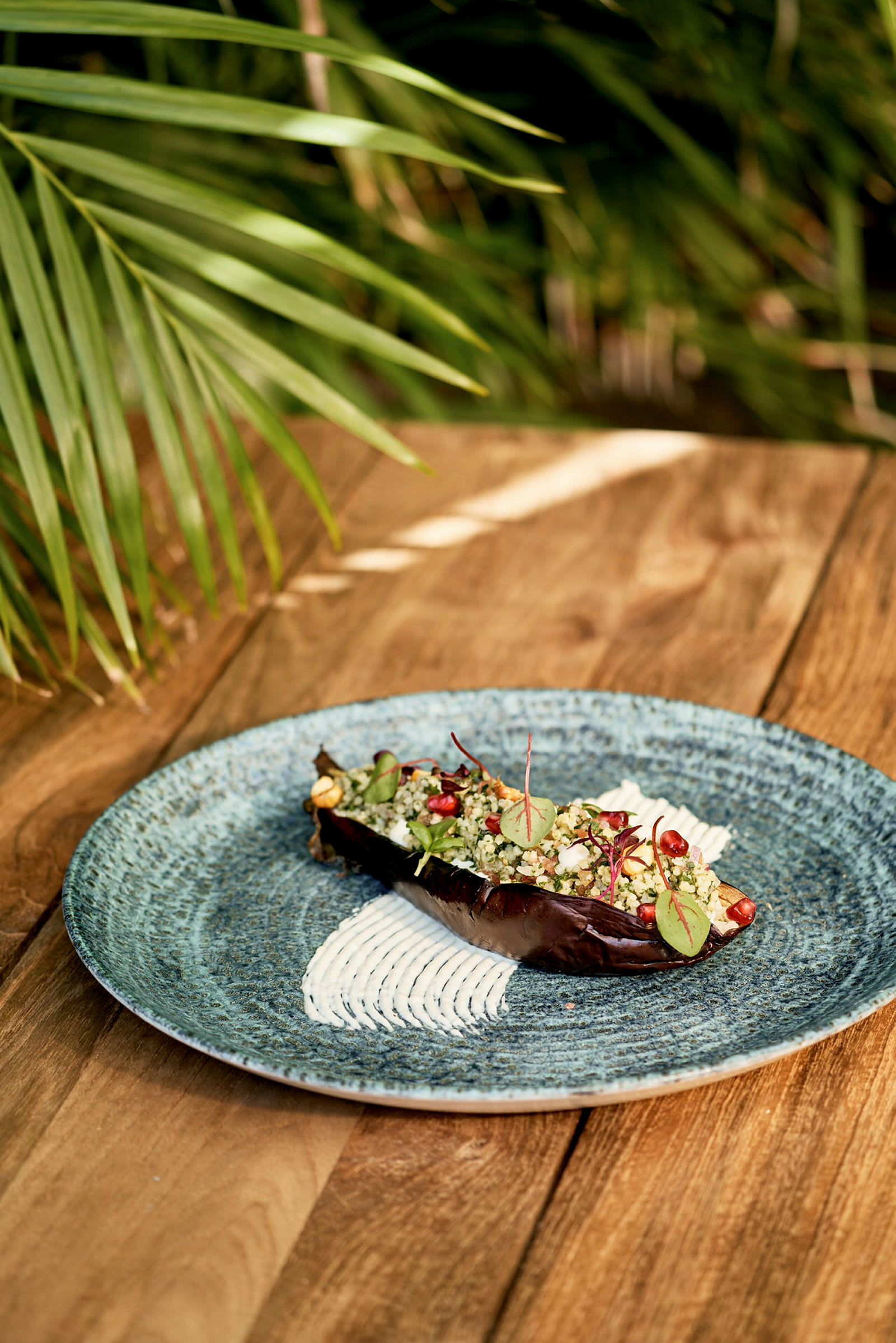 En blå tallerken med en fyldt aubergine, dekoreret med granatæblekerner, urter og creme, står på et træbord. (AI-genereret tekst)