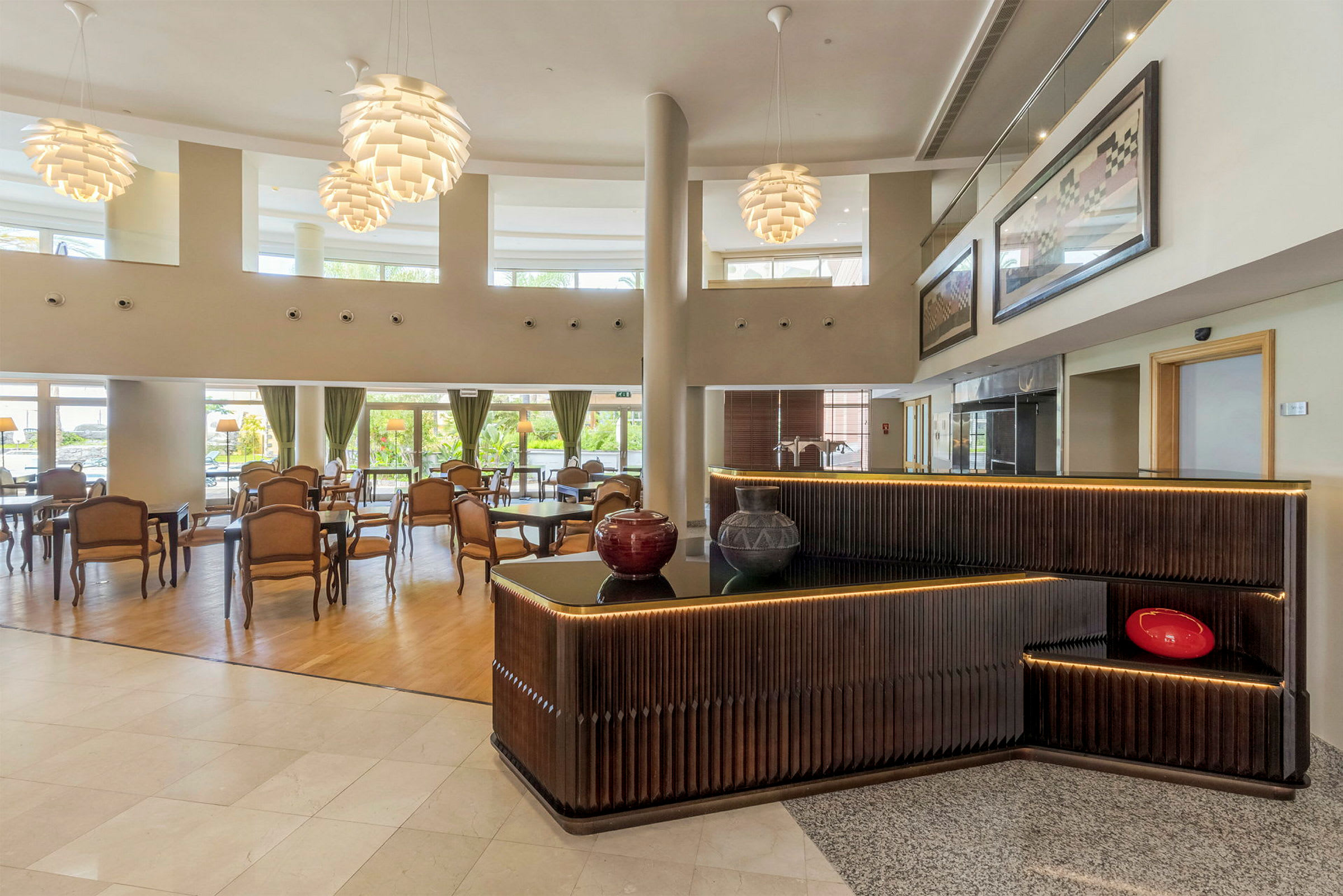 Et elegant hotellobbyområde med borde og stole samt dekorative, moderne lamper. Receptionen har mørkt træpanel og dekorationer. (AI-genereret tekst)