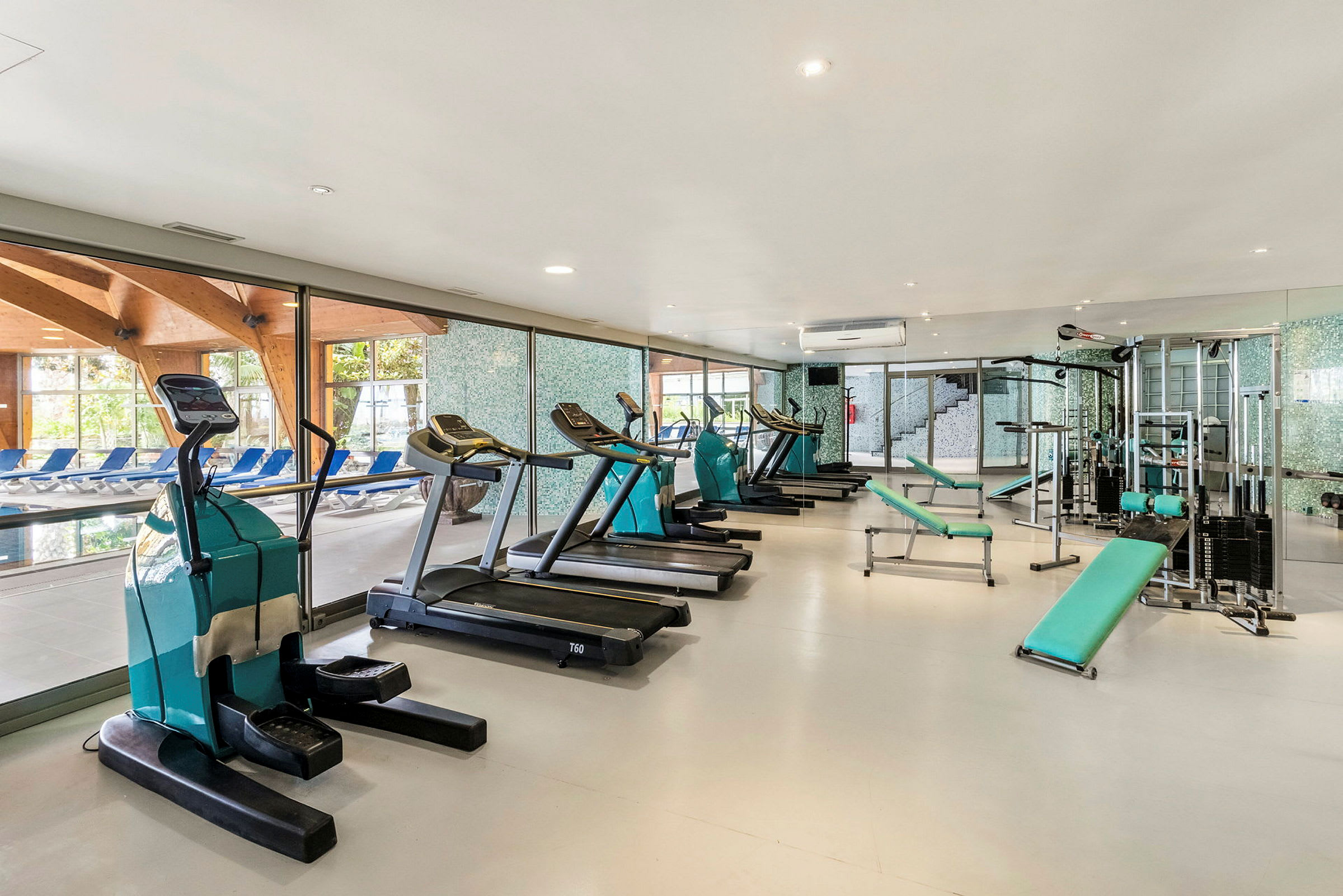 Fitnesscenter med træningsudstyr som crosstrainere, løbebånd og styrketræningsmaskiner, ved siden af store vinduer med udsigt over en pool. (AI-genereret tekst)