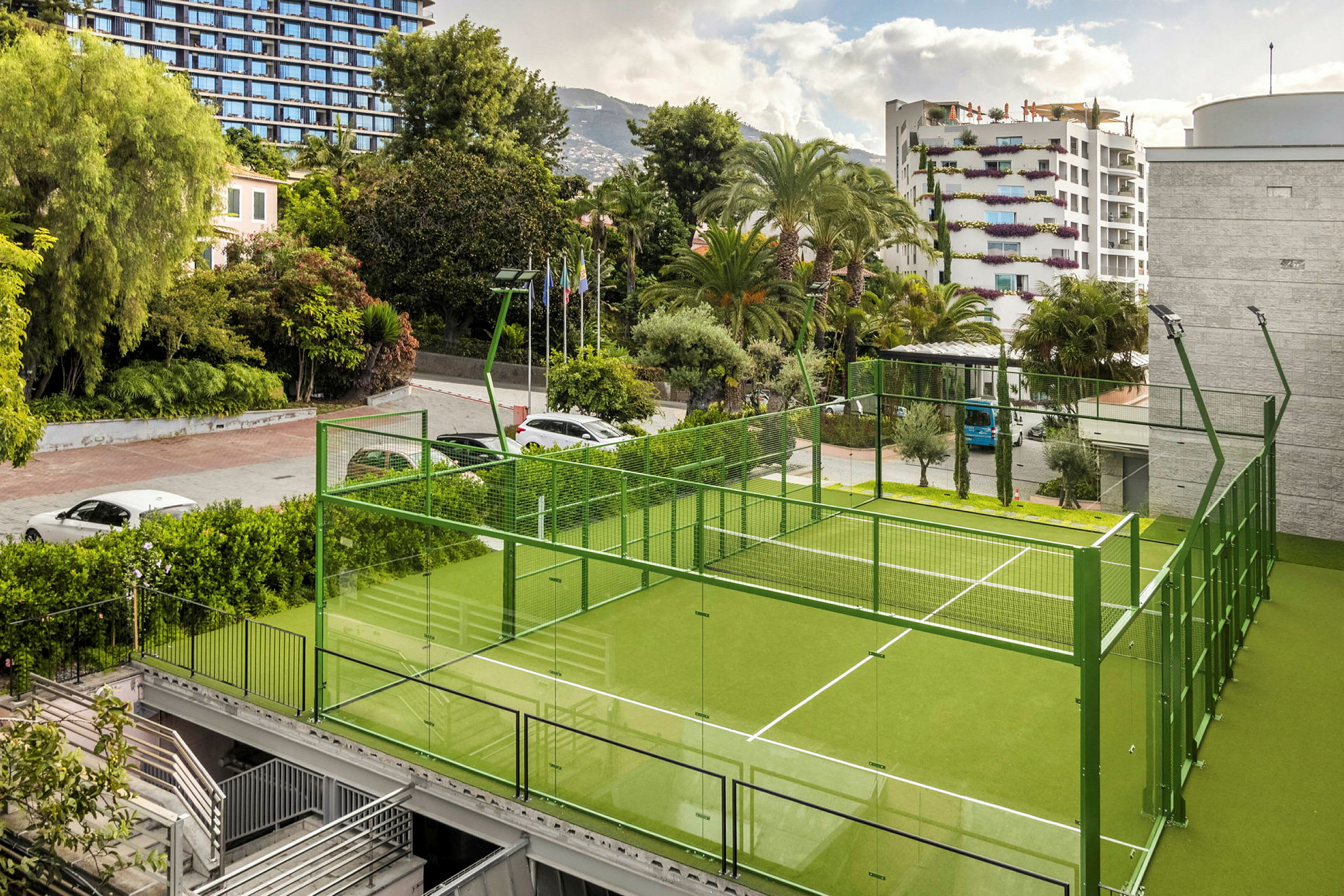 En grøn padelbane med glasvægge omgivet af træer og bygninger. (AI-genereret tekst)