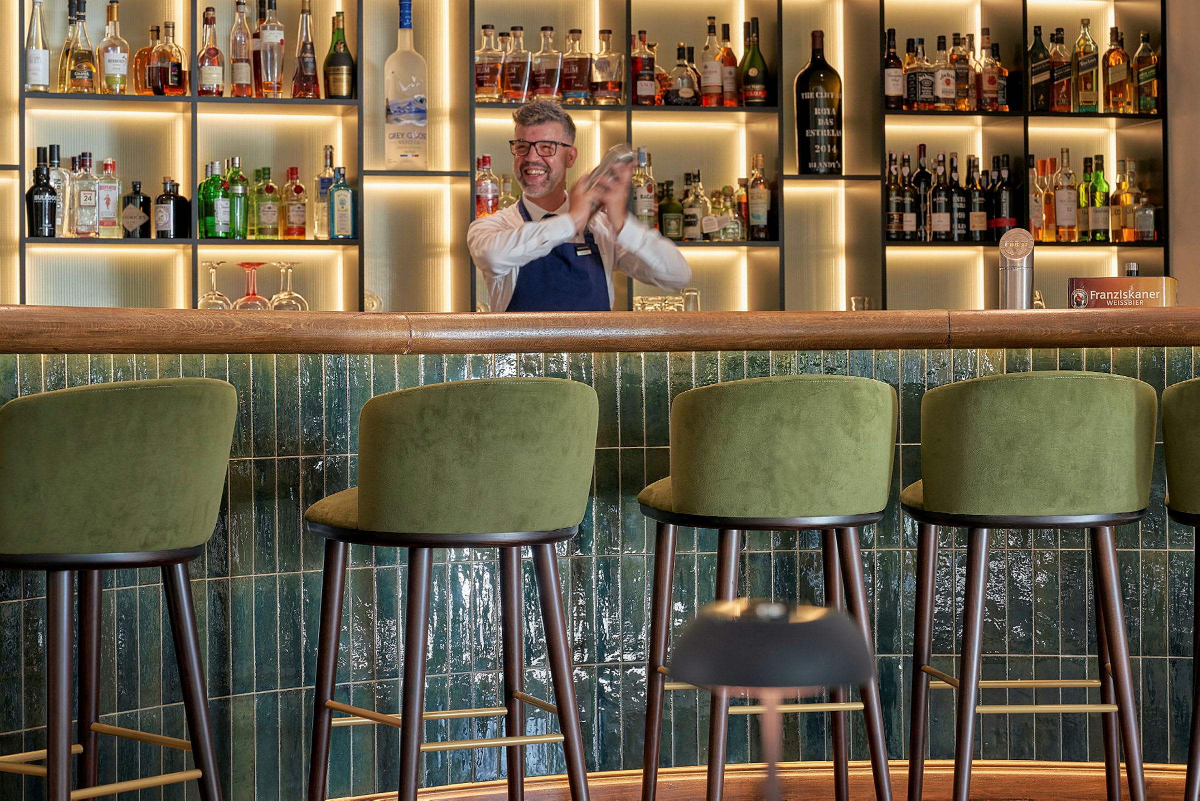En bar med grønne barstole og en hylde med flasker i baggrunden. (AI-genereret tekst)
