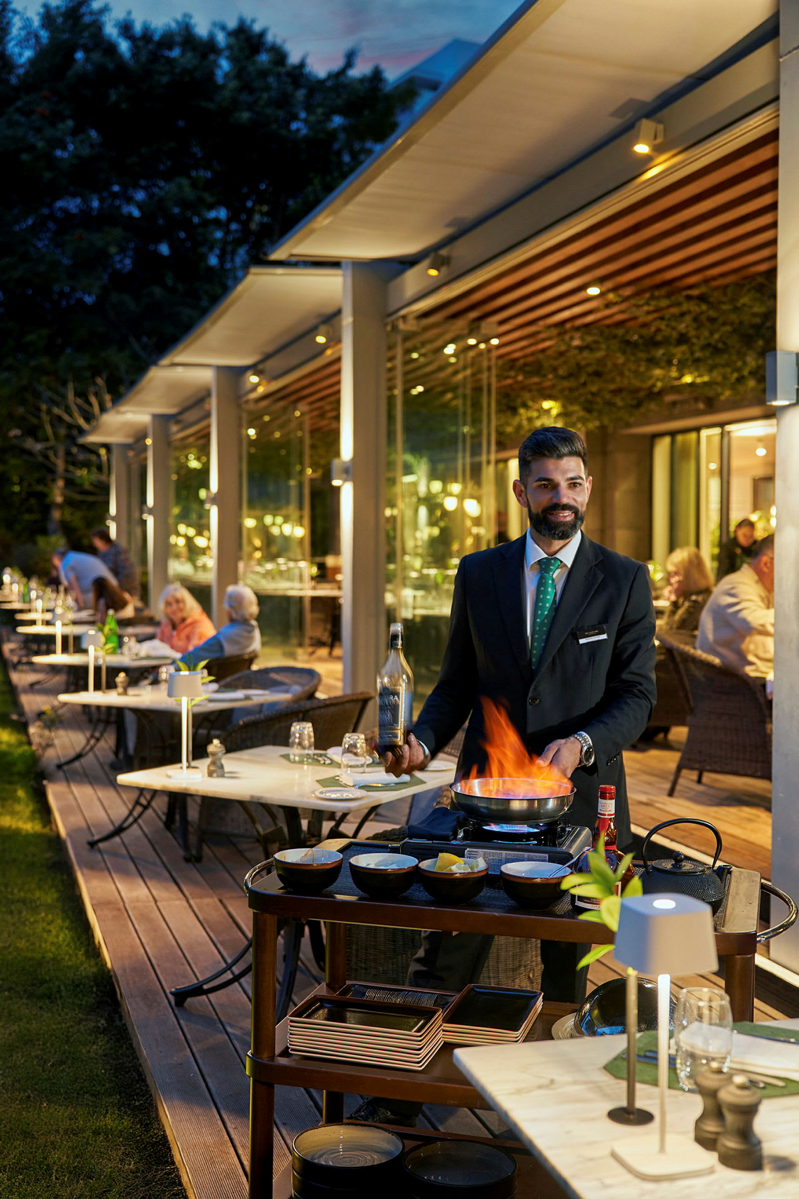 En tjener tilbereder mad ved et bord ved hjælp af en flamberingsbrænder på en restaurant med udendørsservering. (AI-genereret tekst)