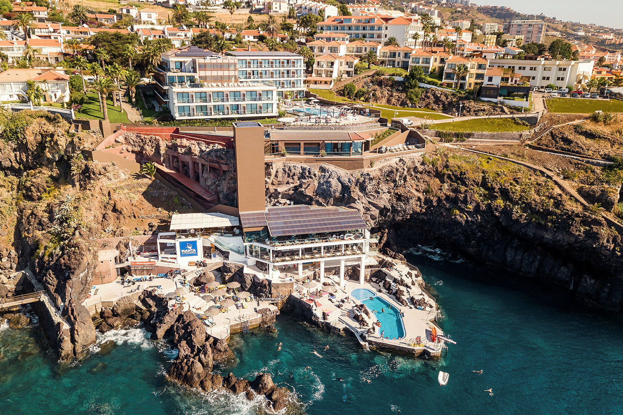 Luftfoto af et hotel beliggende på klipper ved havet, med pool og liggestole. (AI-genereret tekst)