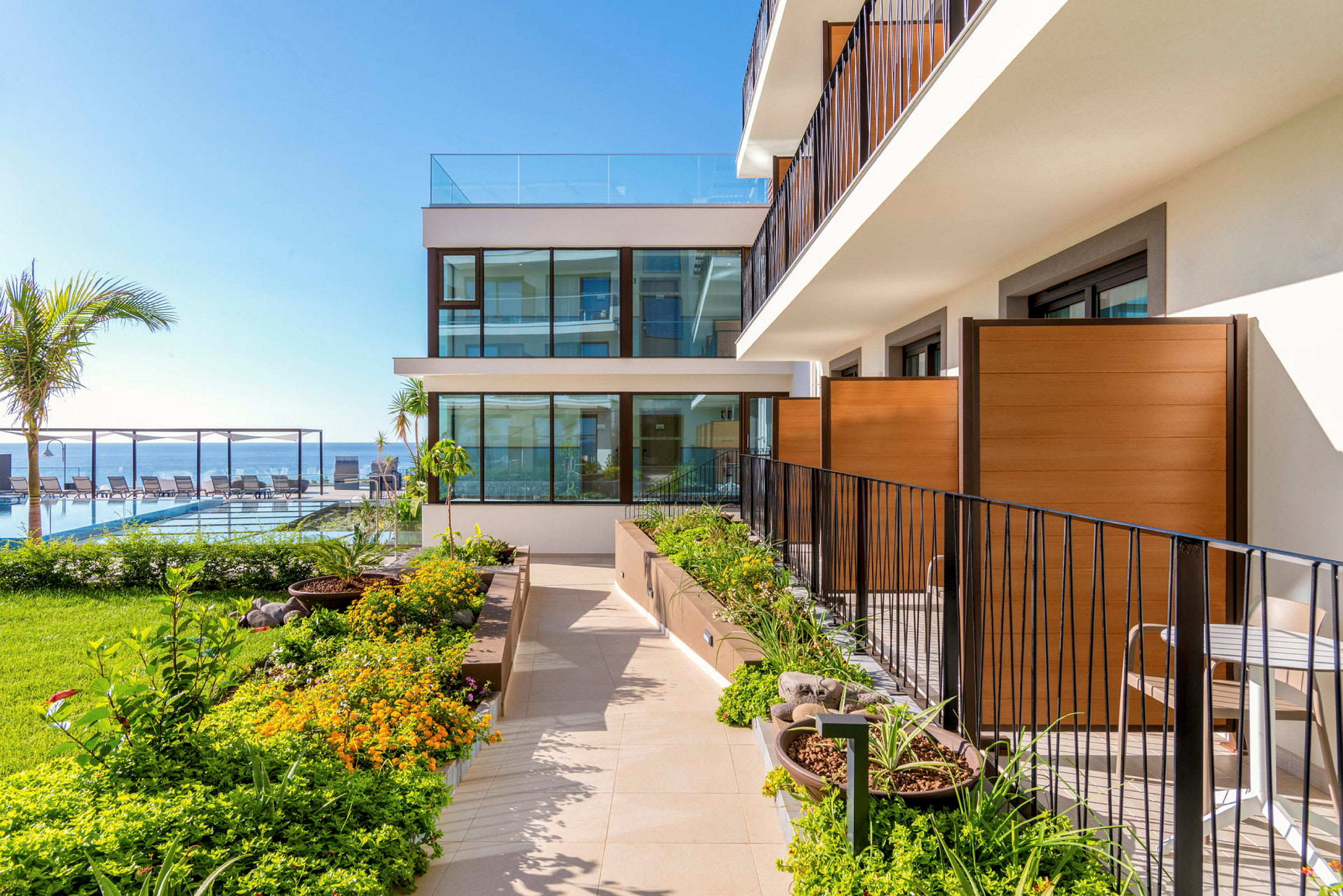Terrasseområde med grønne planter ved en moderne bygning, havet og liggestole er synlige i baggrunden. (AI-genereret tekst)