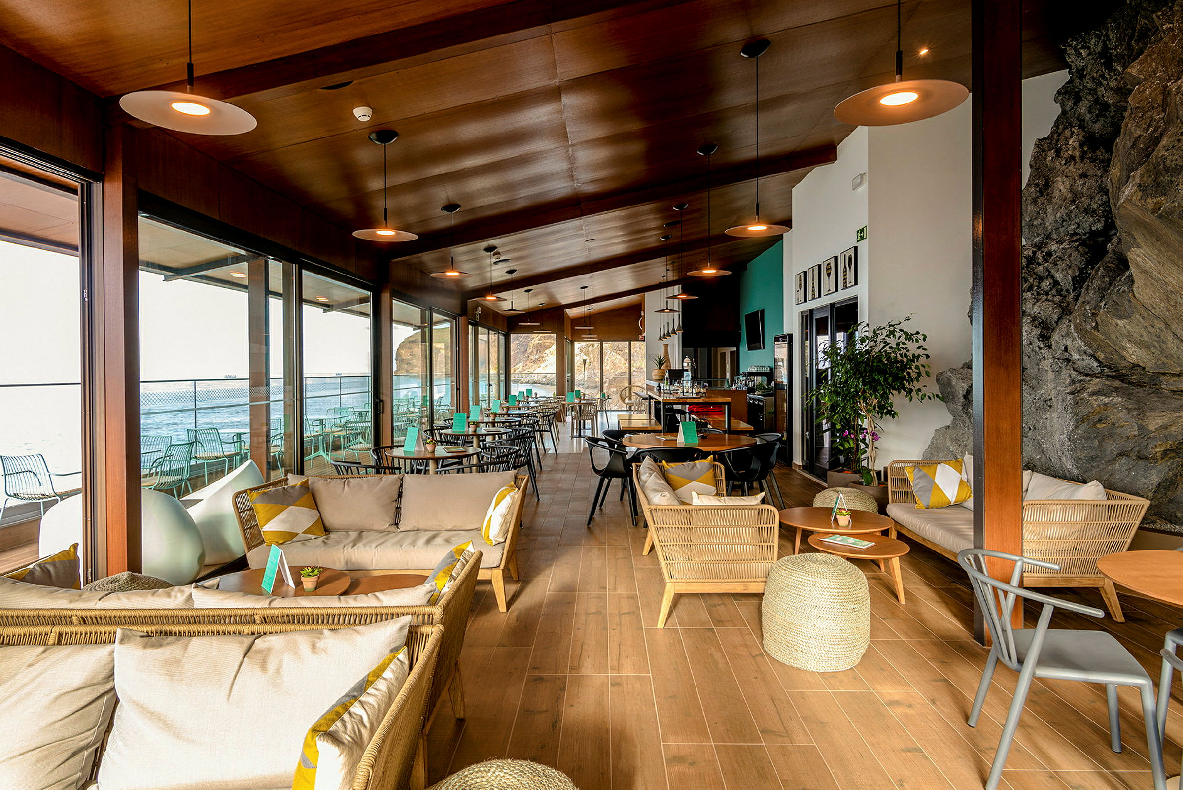 Moderne café med træinteriør, sofaer og borde nær vinduer med udsigt over havet. (AI-genereret tekst)