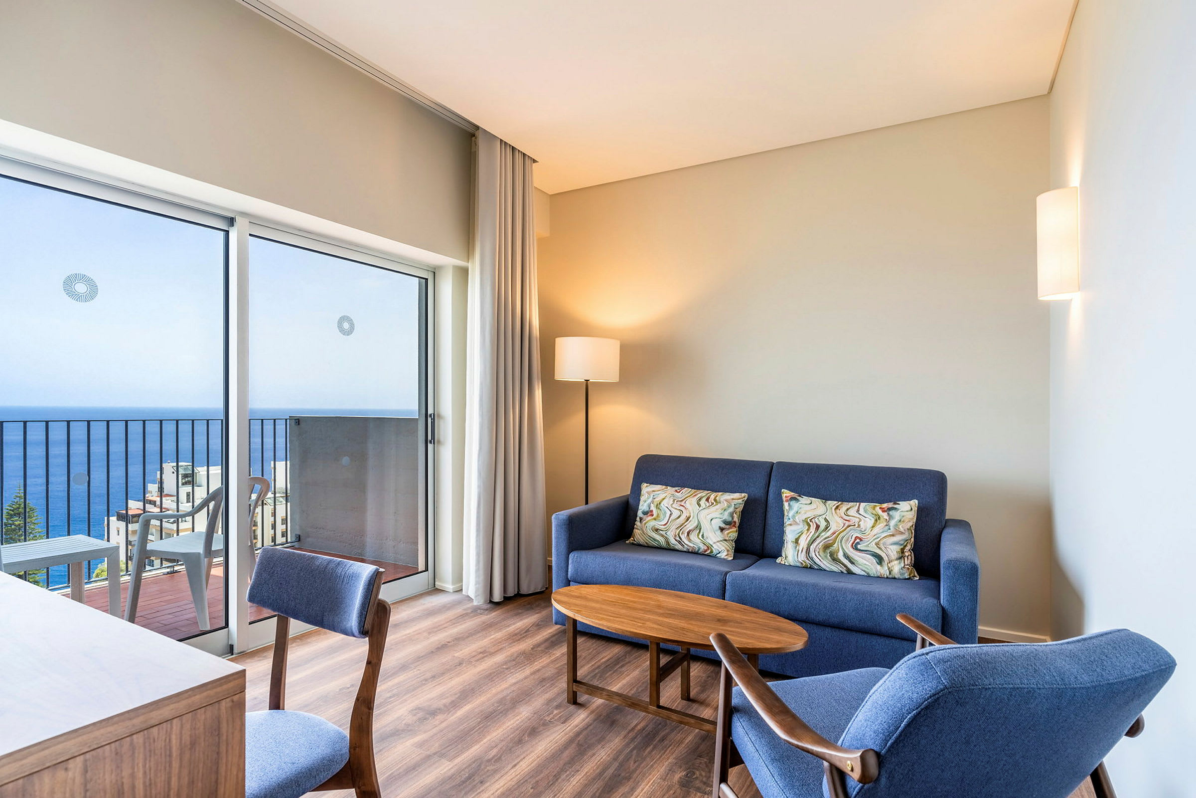 Et moderne indrettet hotelværelse med en blå sofa, blå lænestol, et træbord og en skrivebordsstol. Værelset har en altan med havudsigt. (AI-genereret tekst)