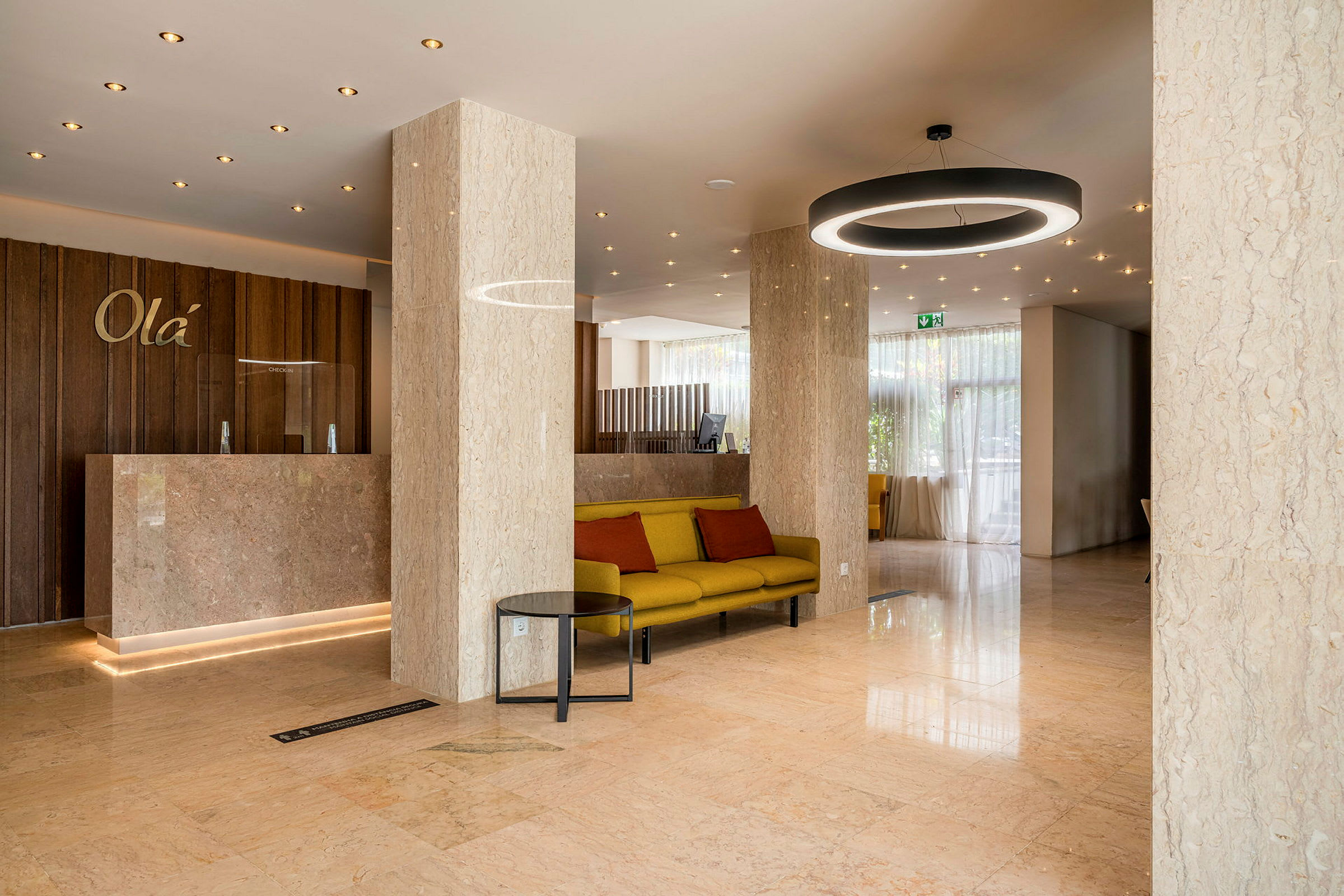 Hotellets lobby med marmorgulv og søjler, en reception med træpanel og en siddegruppe med gul sofa og røde puder. (AI-genereret tekst)