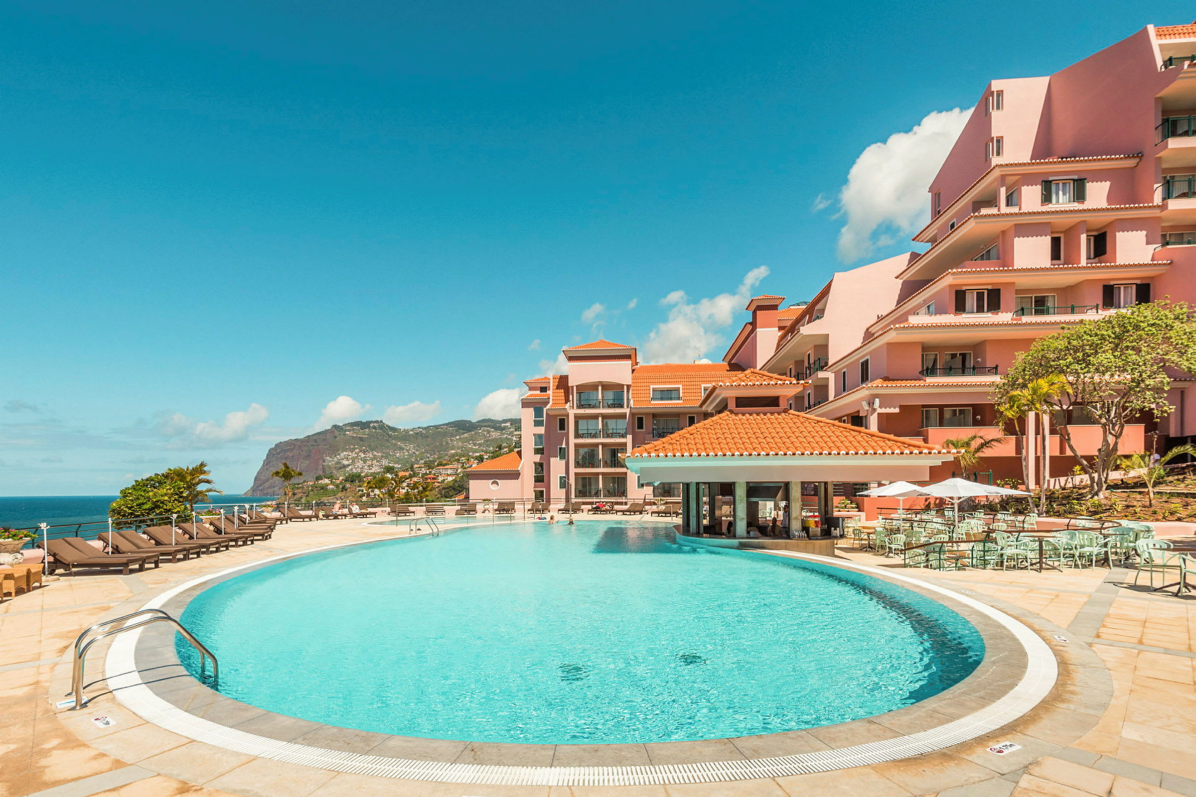 En hotelpool omgivet af solsenge og lyserøde bygninger med havudsigt. (AI-genereret tekst)