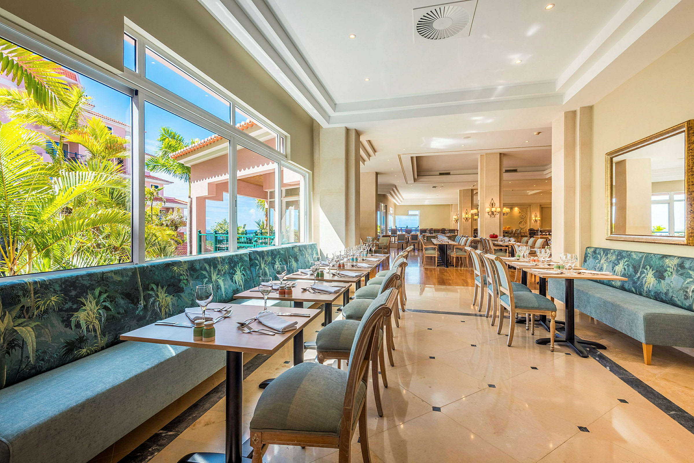 En lys restaurant med moderne borde og stole samt store vinduer med udsigt til grønne omgivelser. (AI-genereret tekst)