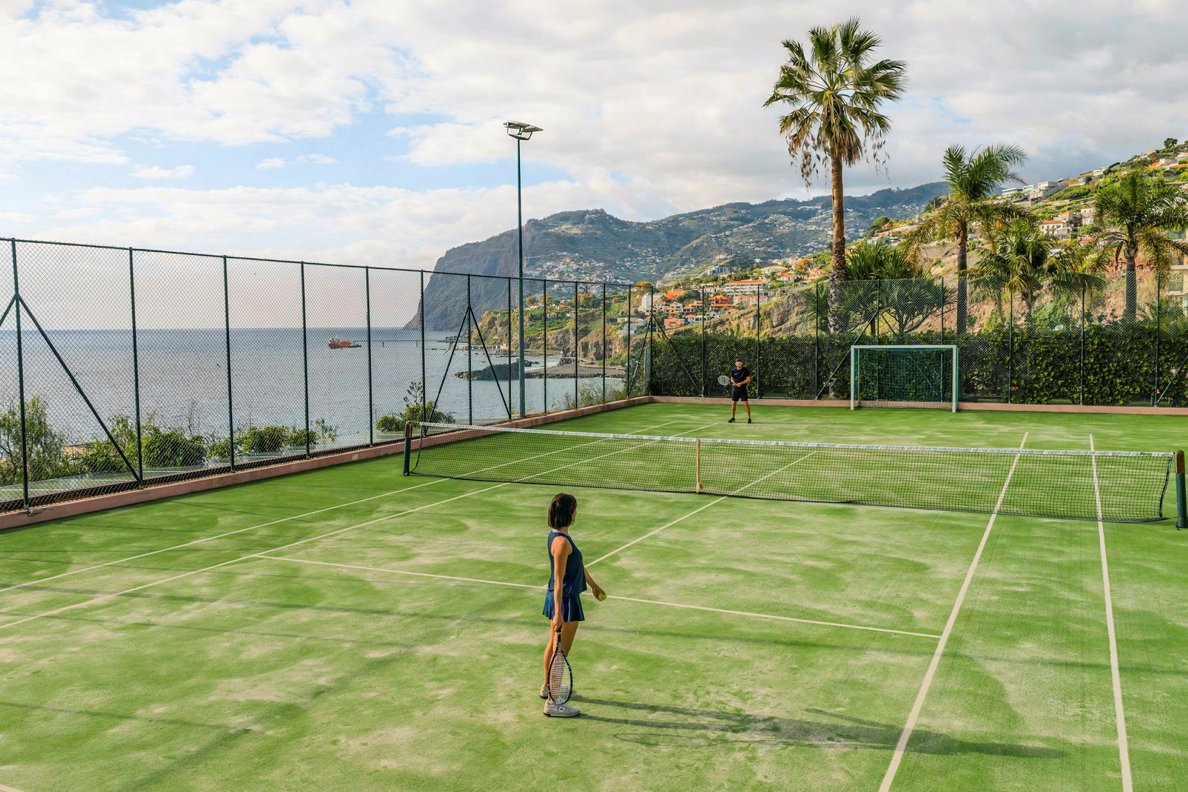 To personer spiller tennis på en udendørsbane med udsigt over havet og omkringliggende bjerge. (AI-genereret tekst)
