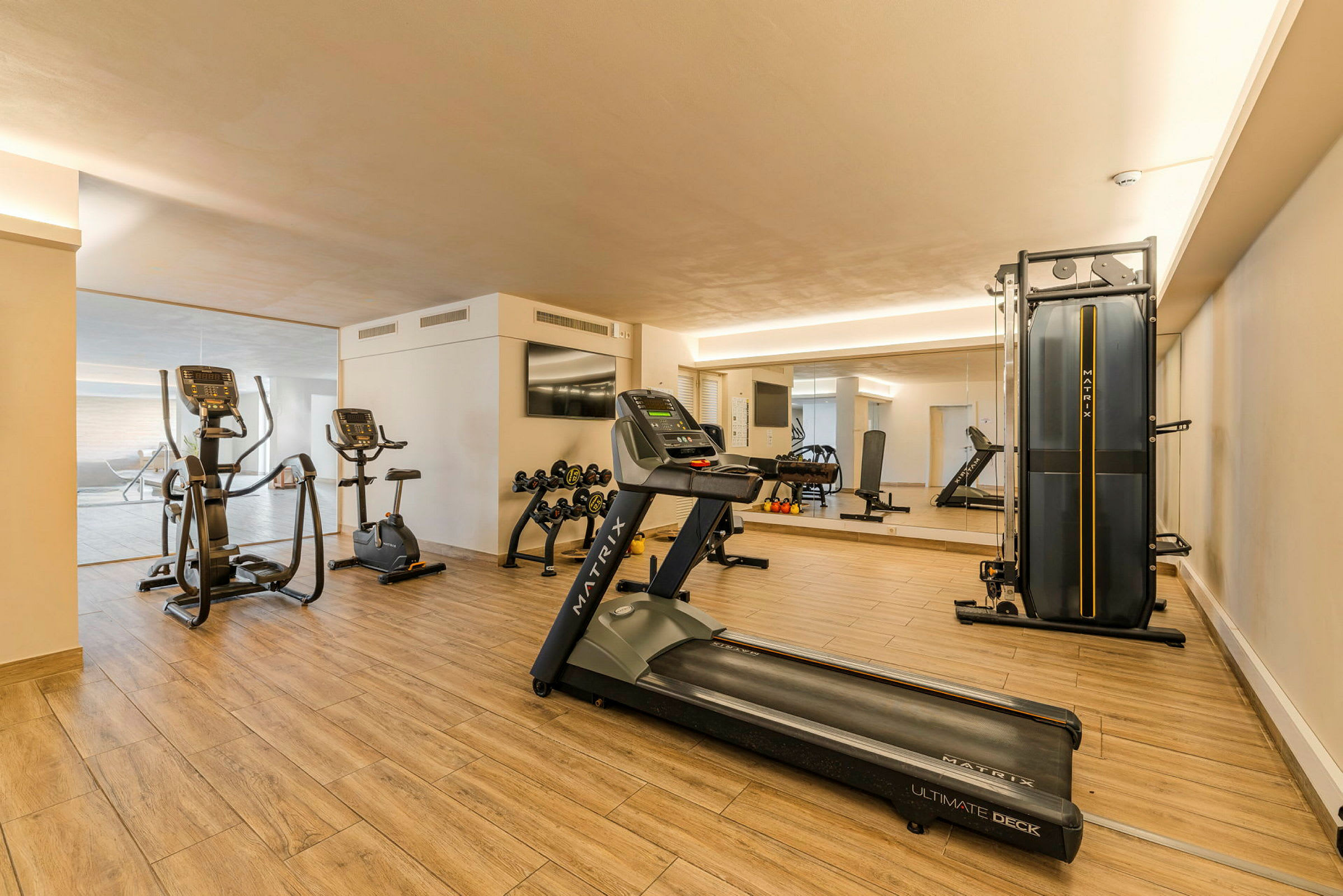 Et fitnessrum med træningsmaskiner, herunder løbebånd, motionscykler og vægte. Rummet har trægulv og et stort spejl. (AI-genereret tekst)