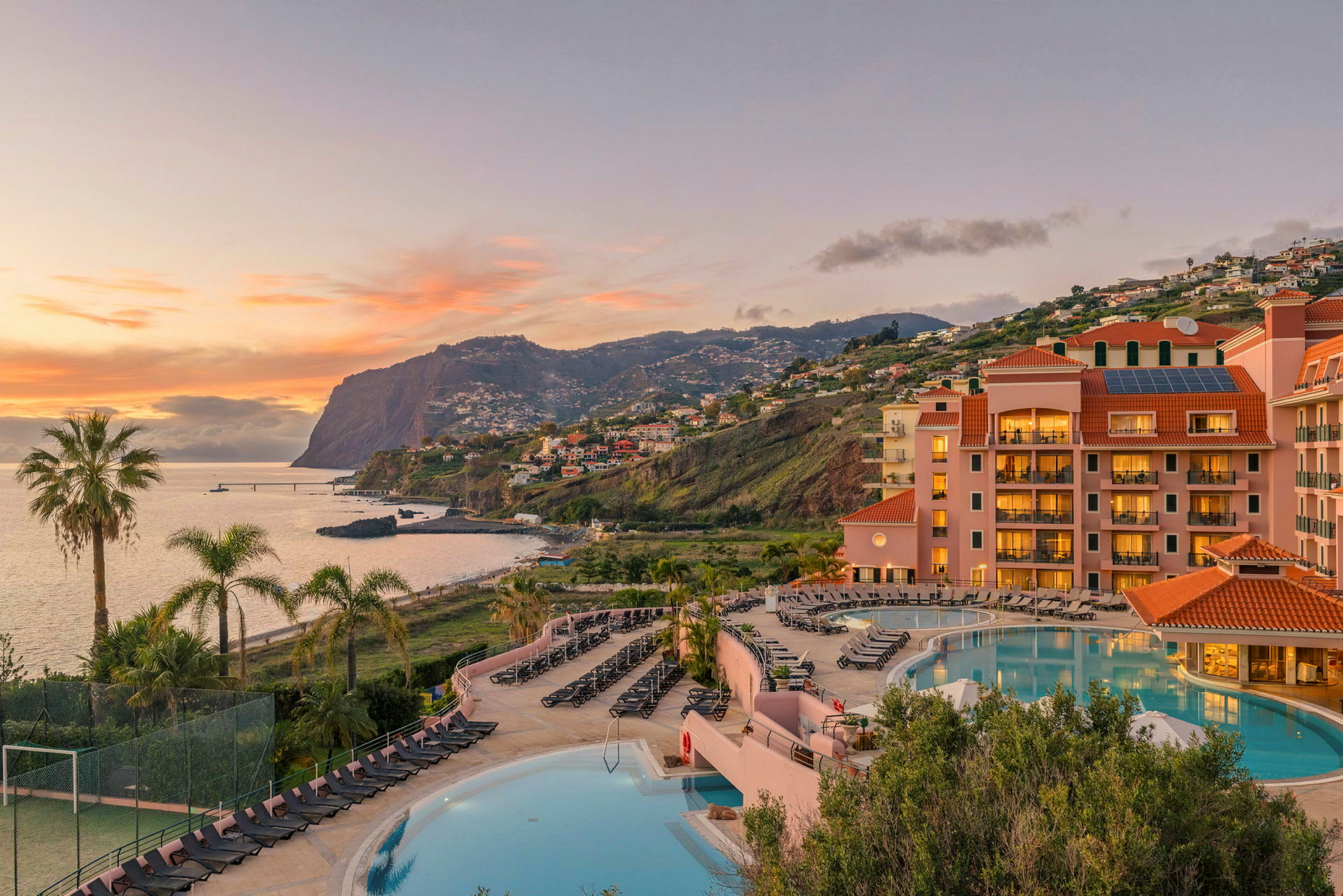 Et hotel med en swimmingpool i forgrunden og en fantastisk udsigt over havet og bjergene i baggrunden. (AI-genereret tekst)