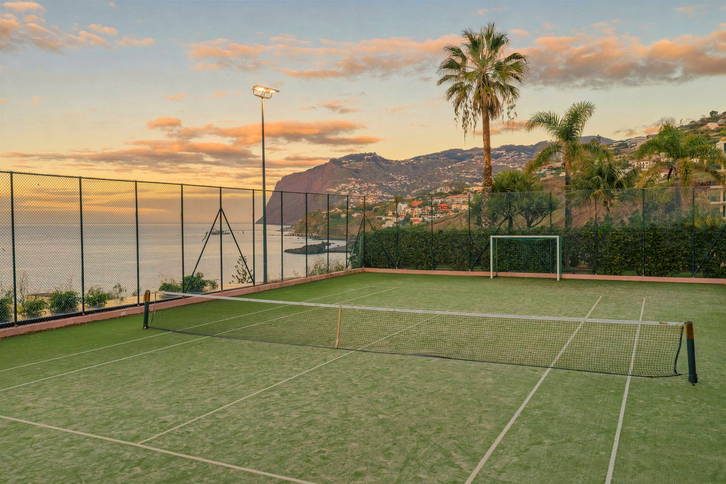 En tennisbane nær havet, omgivet af hegn, palmer og bjerge i baggrunden. (AI-genereret tekst)