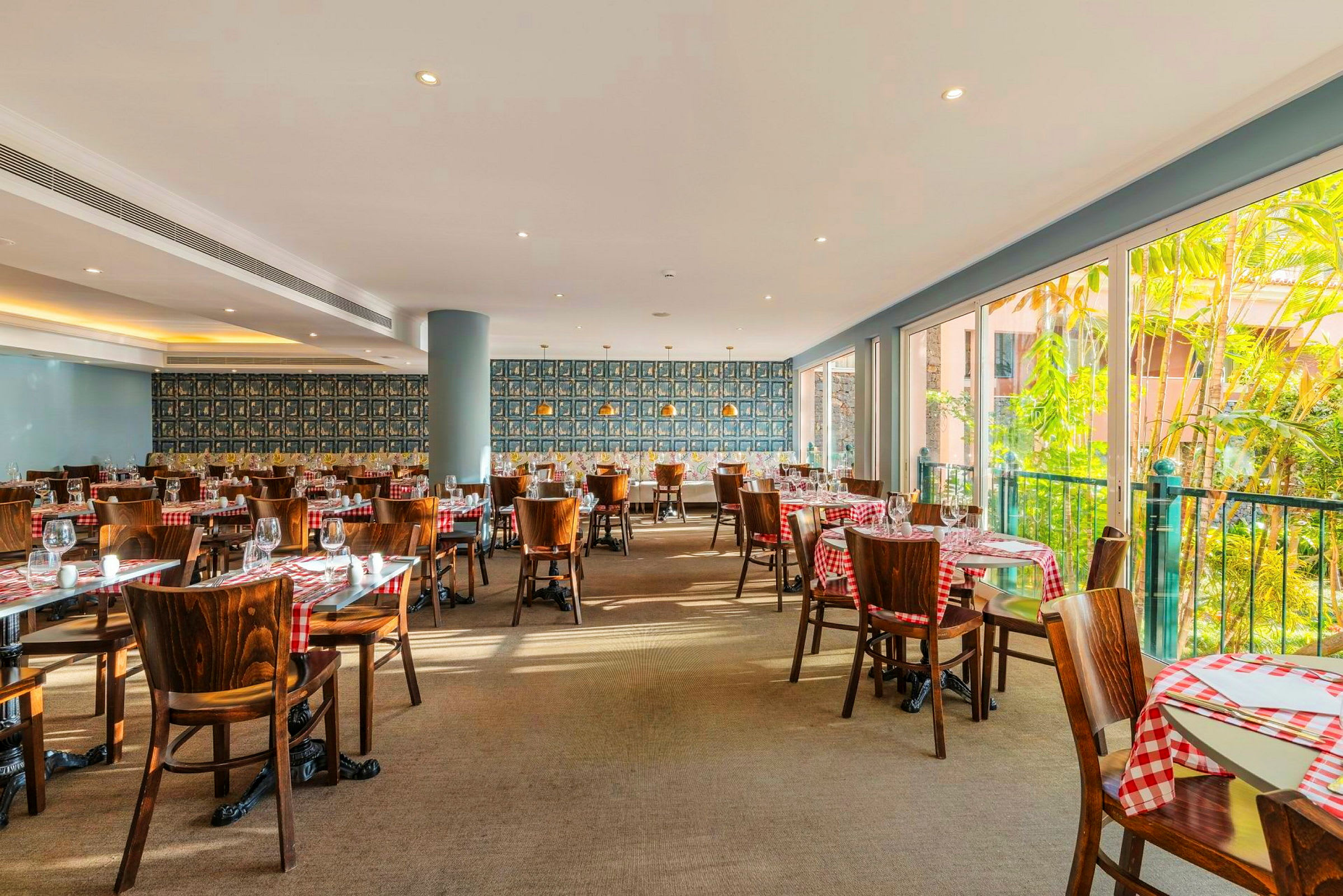 En lyst indrettet restaurant med rød- og hvidternede duge på træborde og stole. Store vinduer lader dagslyset strømme ind og afslører grønne planter udenfor. (AI-genereret tekst)
