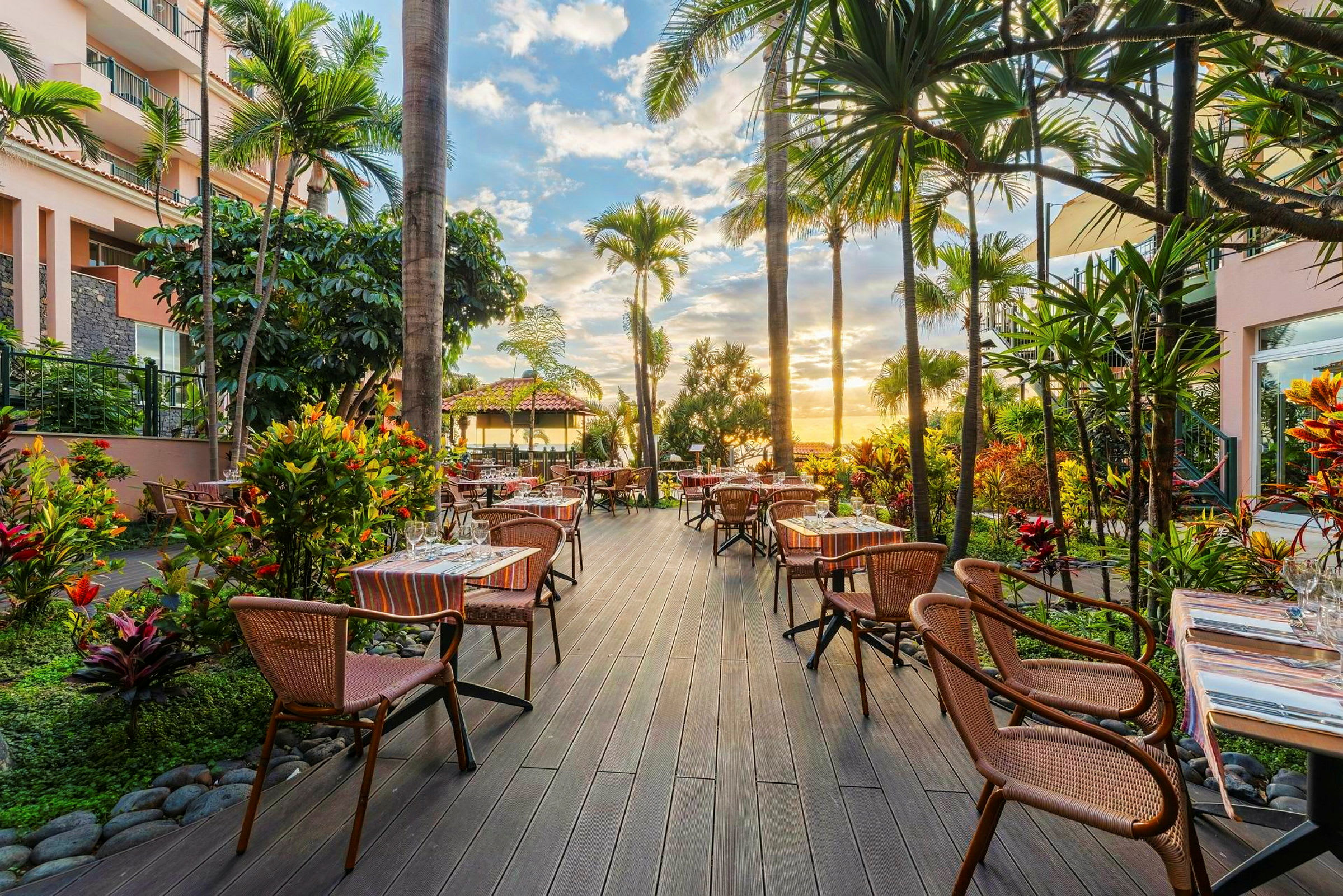 En udendørsrestaurant omgivet af grønne planter og palmer med borde og stole arrangeret på en terrasse, og en solnedgang er synlig i baggrunden. (AI-genereret tekst)