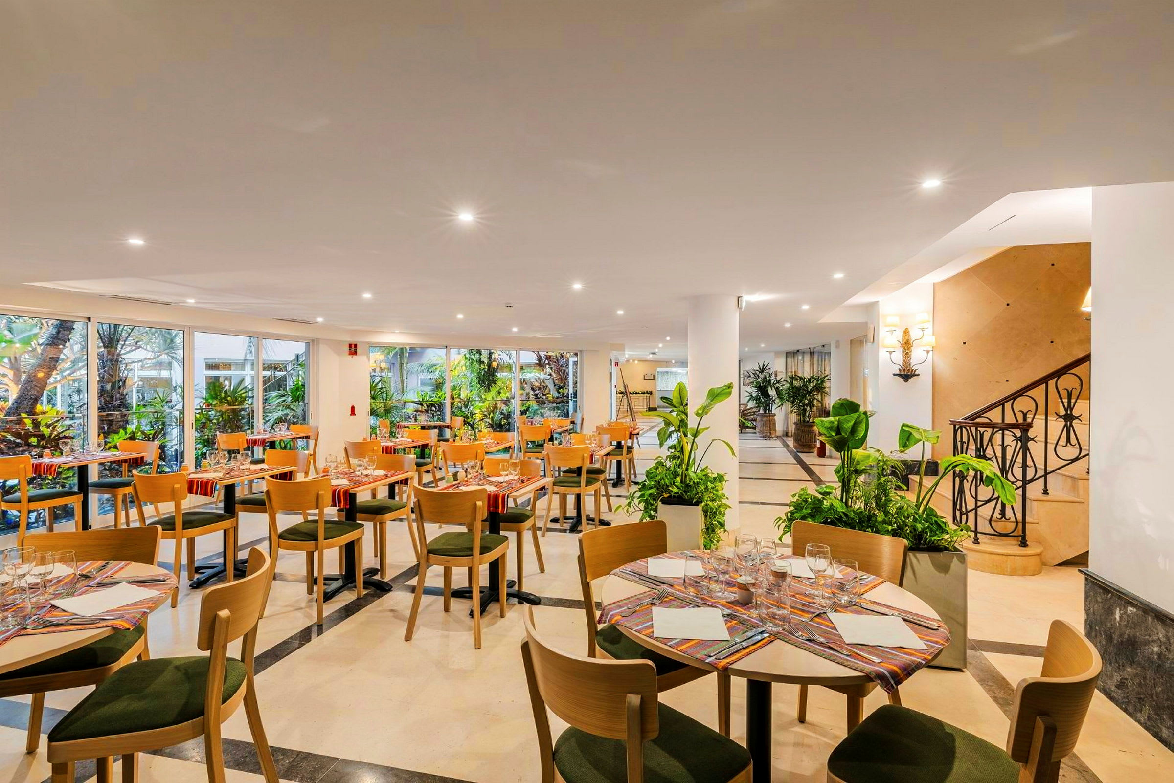 En veloplyst restaurant med flere dækkede borde omgivet af glasvinduer og planter. (AI-genereret tekst)