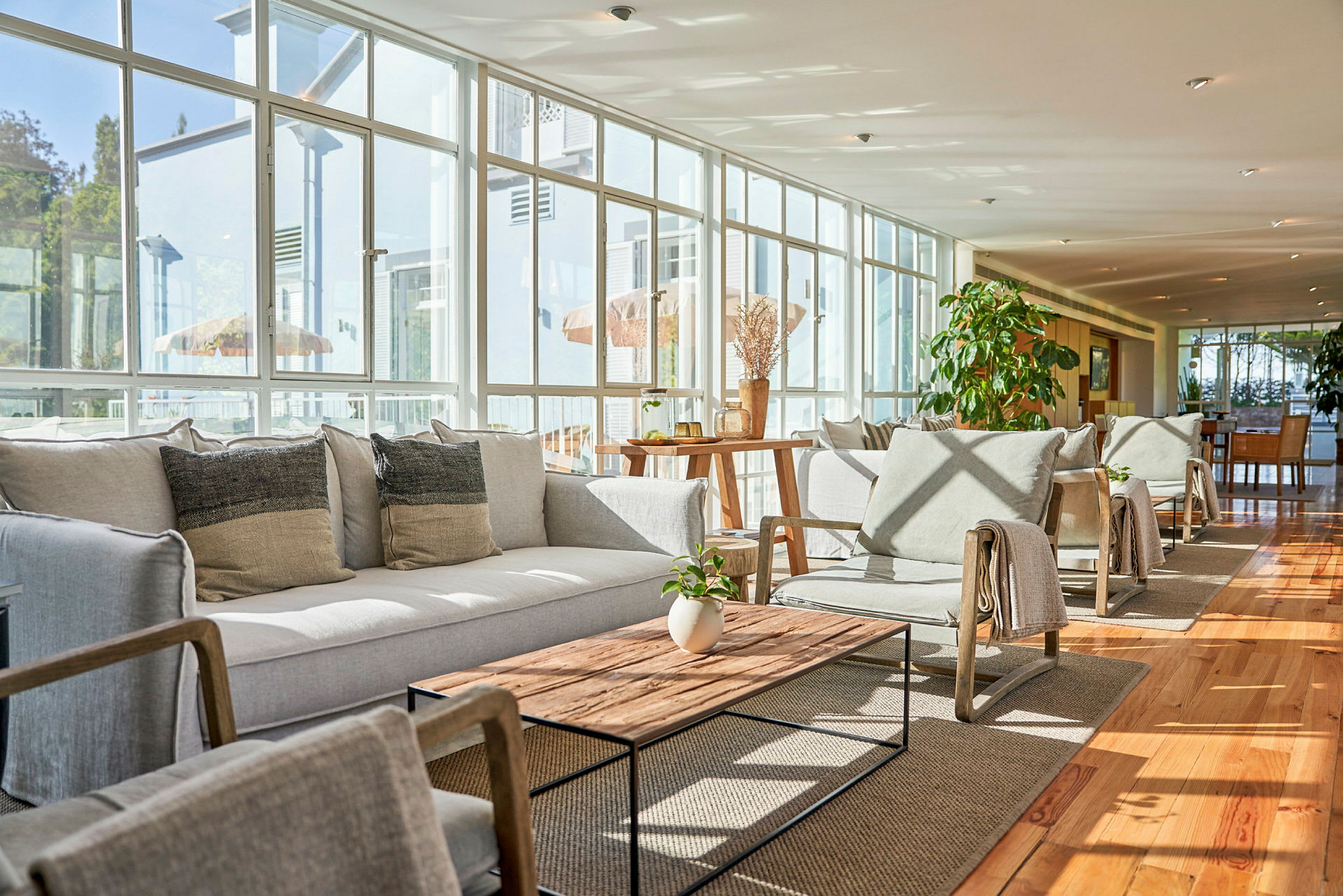 Lys lounge med siddepladser og sofaborde, omgivet af store vinduer. (AI-genereret tekst)