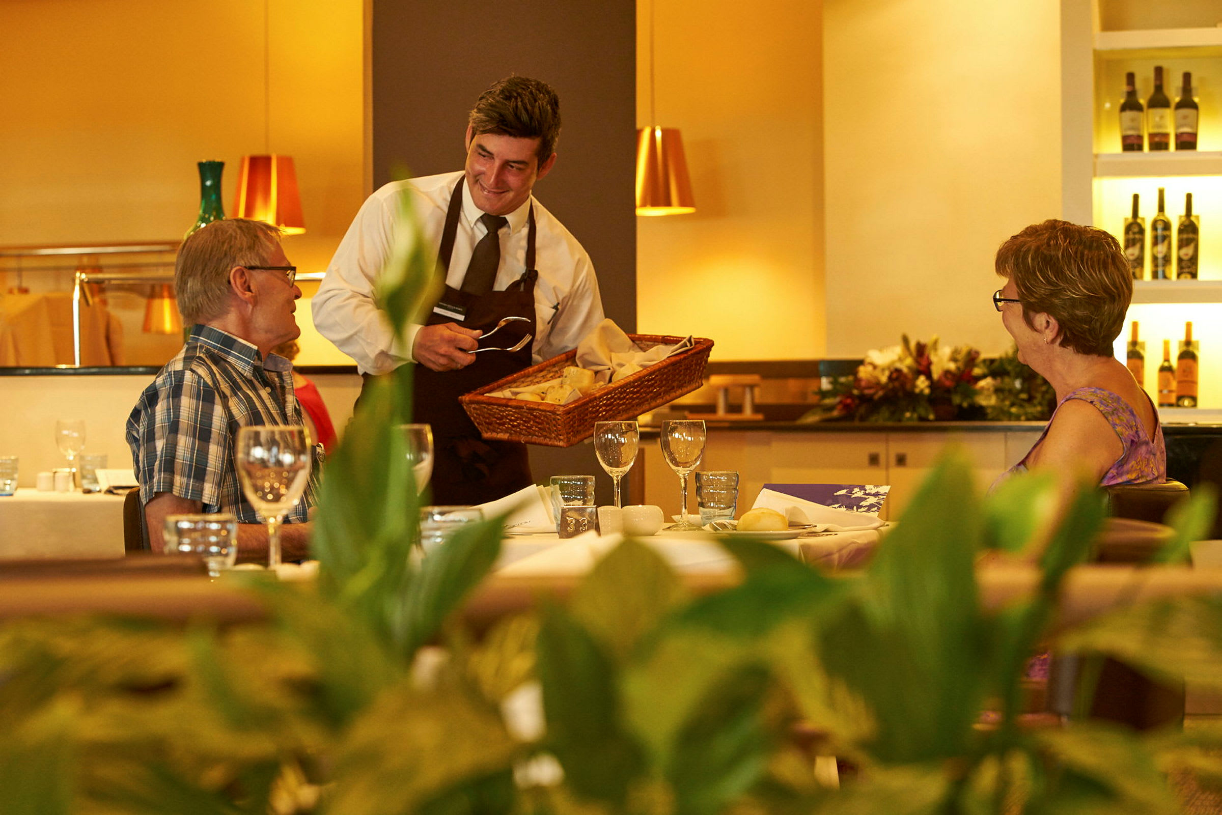 En tjener tilbyder brød til gæster ved et dækket bord i en restaurant. (AI-genereret tekst)
