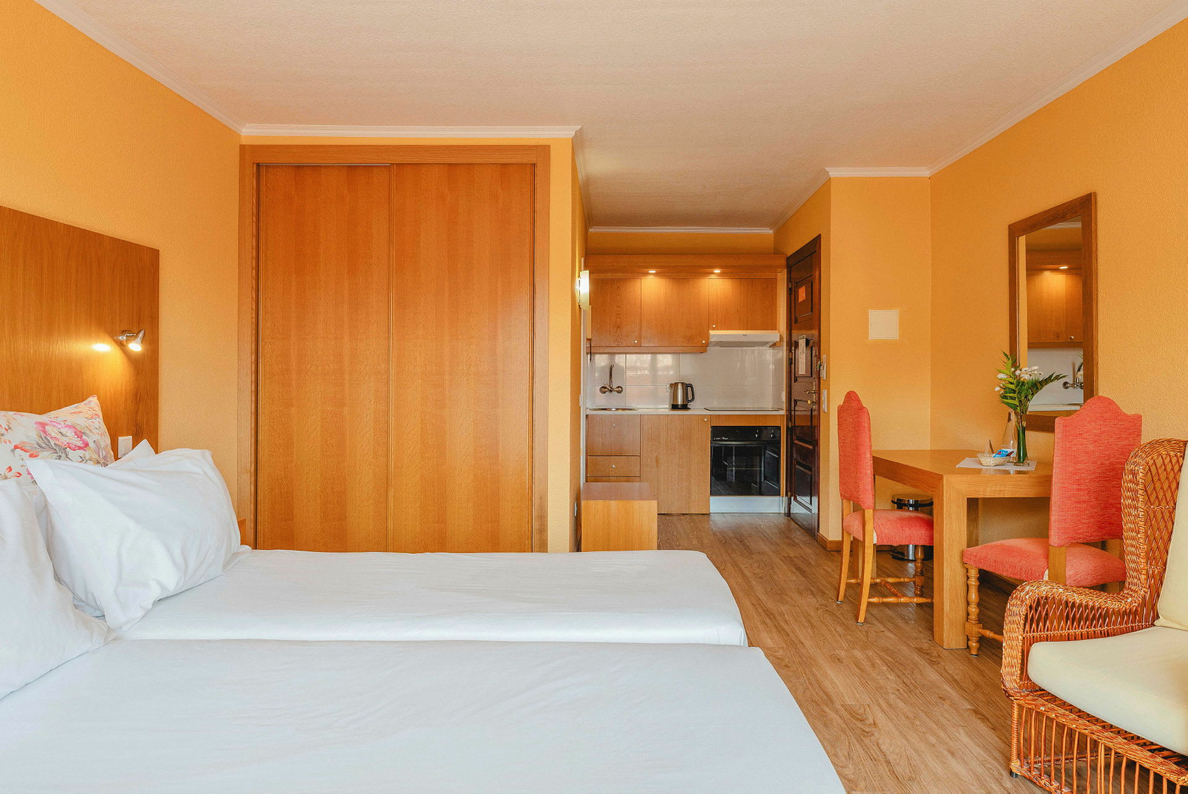 Hotelværelse med to enkeltsenge, træmøbler, orange vægge og et lille køkken i baggrunden. (AI-genereret tekst)