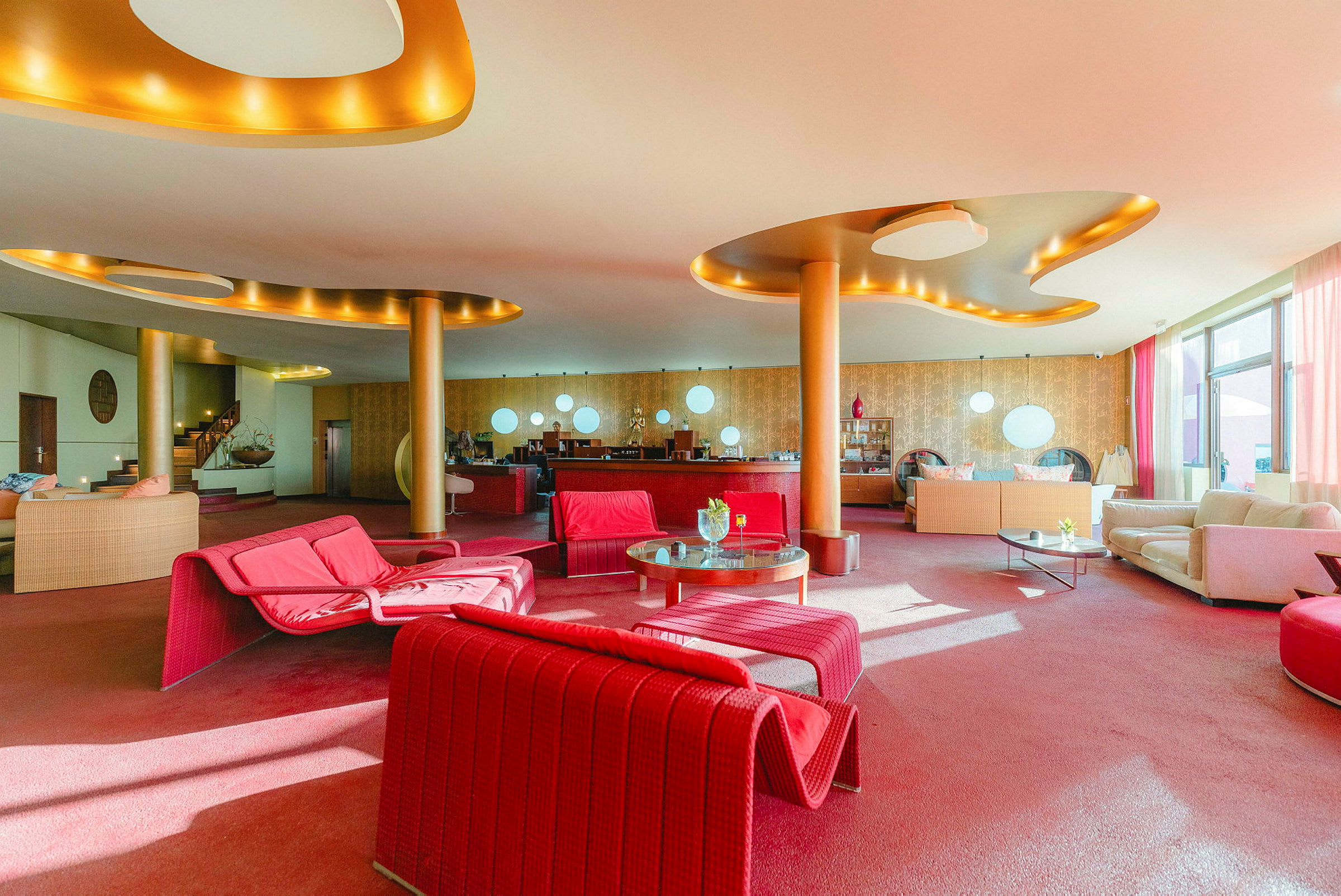 En hotellobby med farverige møbler, primært røde og beige. Der findes sofaer, borde og moderne indretningsdetaljer. (AI-genereret tekst)