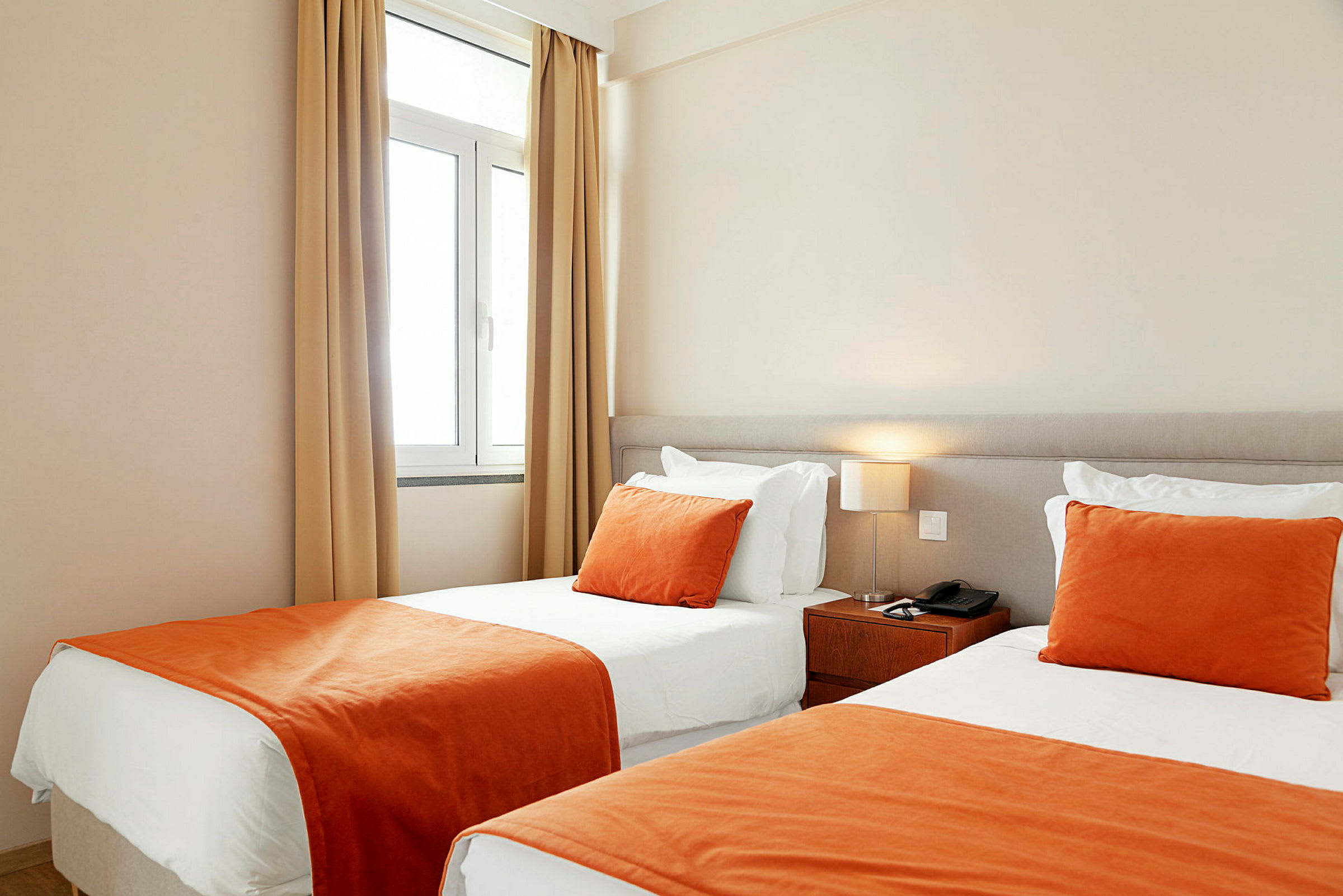 Et hotelværelse med to enkeltsenge ved siden af hinanden, med hvide lagner og orange tæpper og pyntepuder. (AI-genereret tekst)