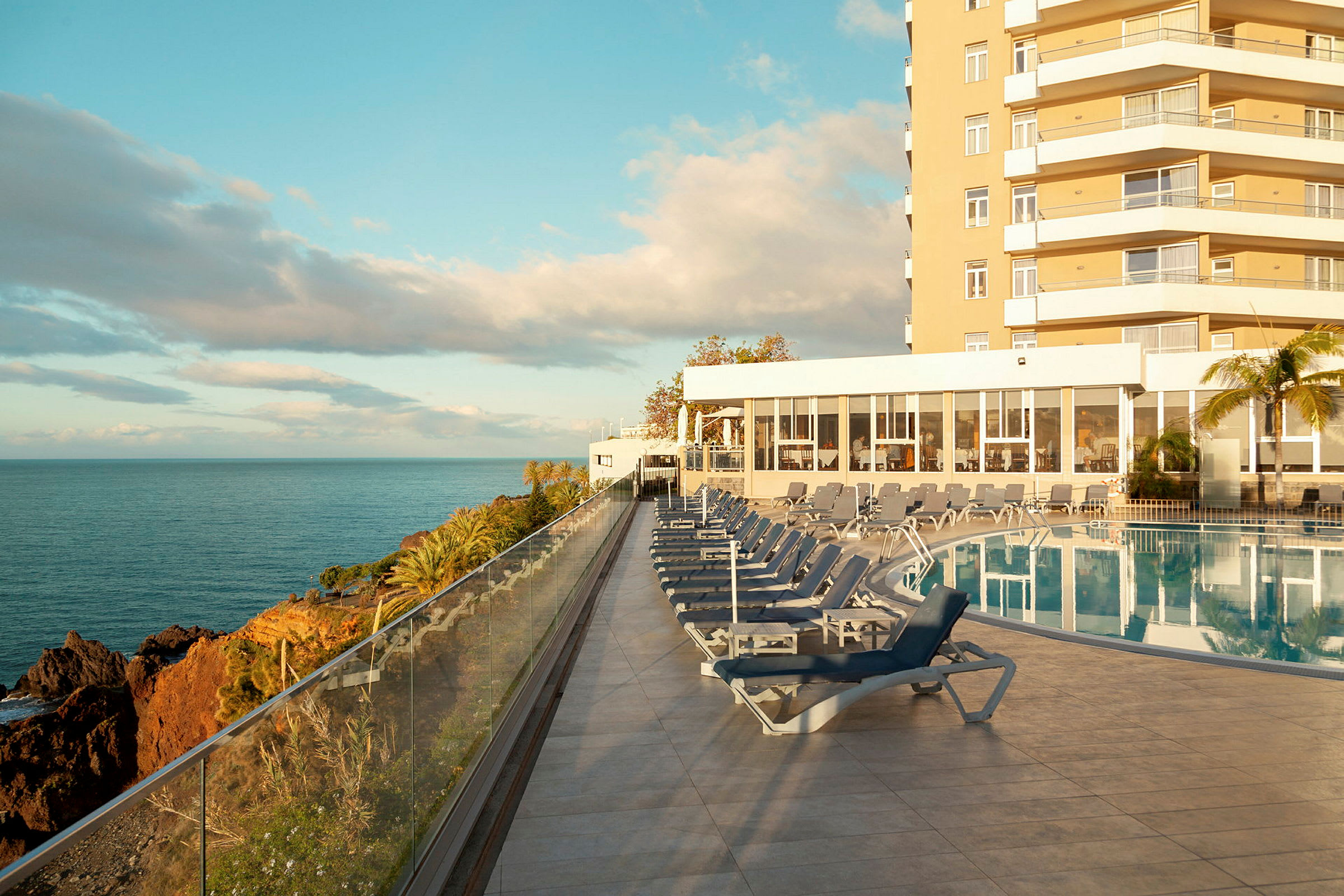 Udendørs pool med liggestole, havudsigt og hotelbygningen i baggrunden. (AI-genereret tekst)