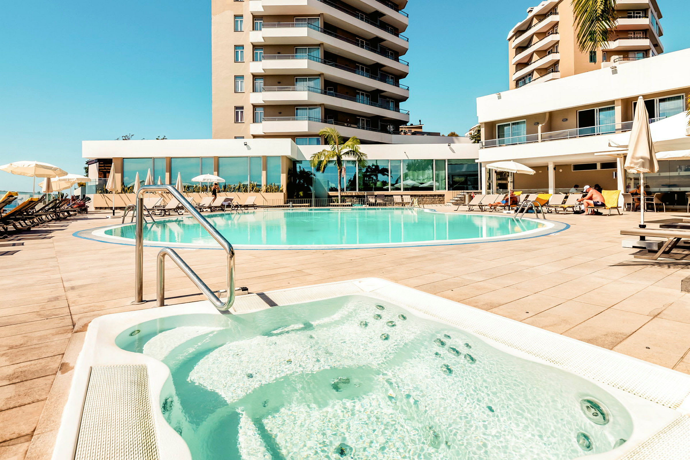 Billede af et hotelpoolområde med et boblebad i forgrunden, liggestole og parasoller samt omkringliggende bygninger. (AI-genereret tekst)