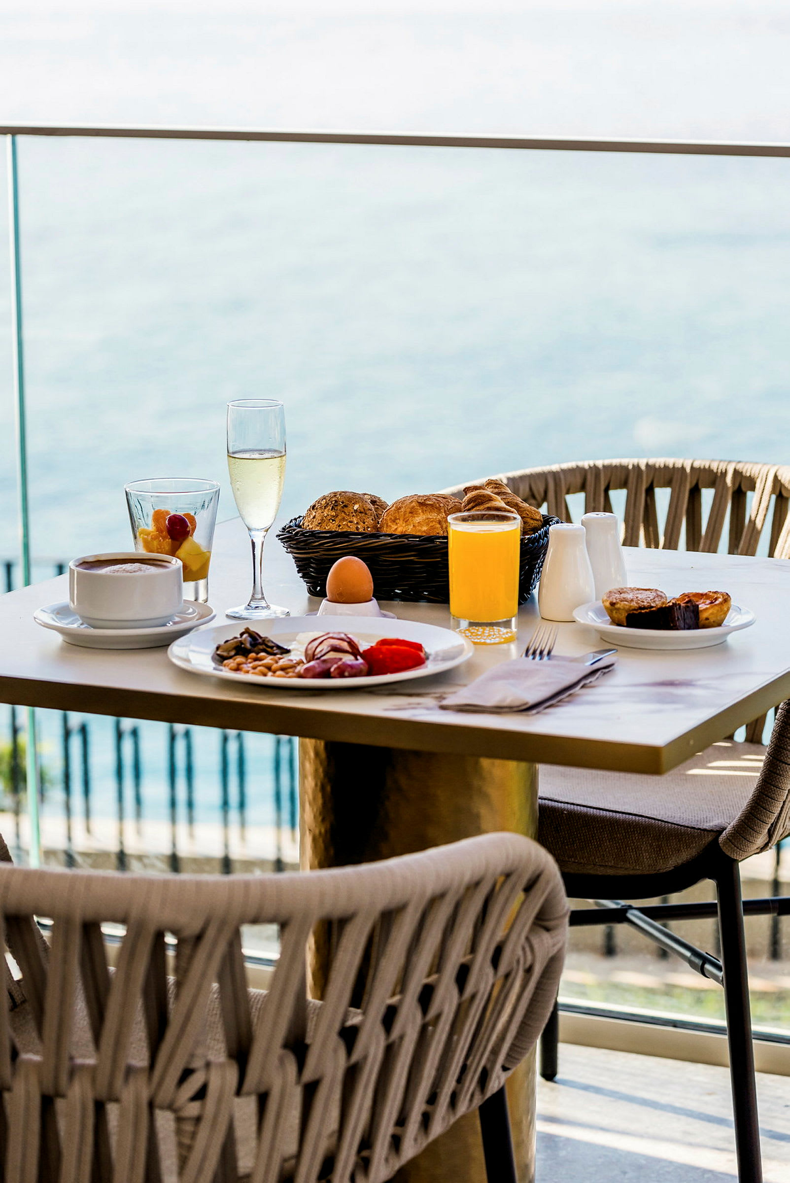 Morgenbord med forskellige retter, drikkevarer og brød, med udsigt til havet i baggrunden. (AI-genereret tekst)
